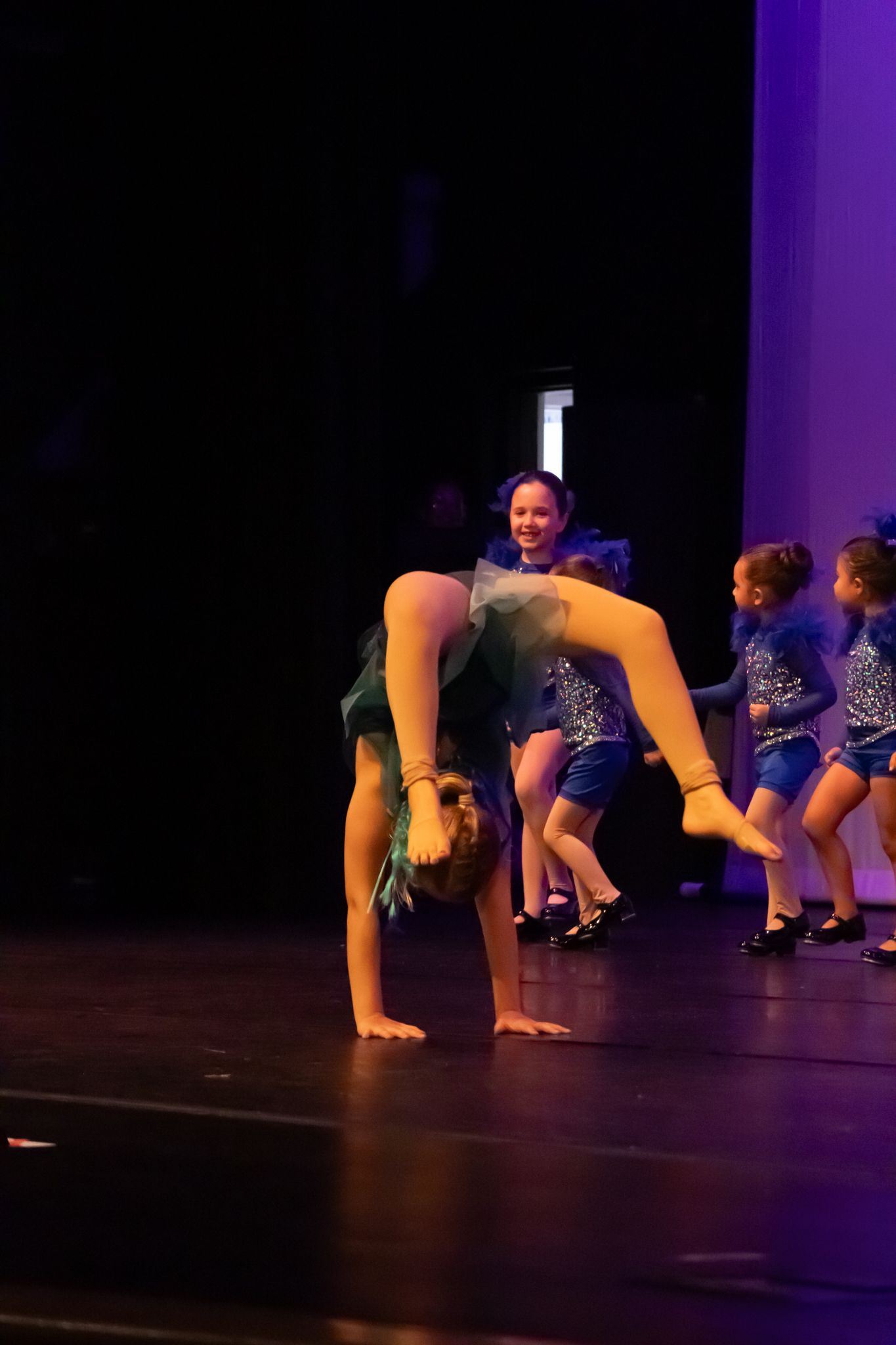 Young dancers perform acrobatic moves on stage, with one girl doing a backbend in a teal sequined costume.