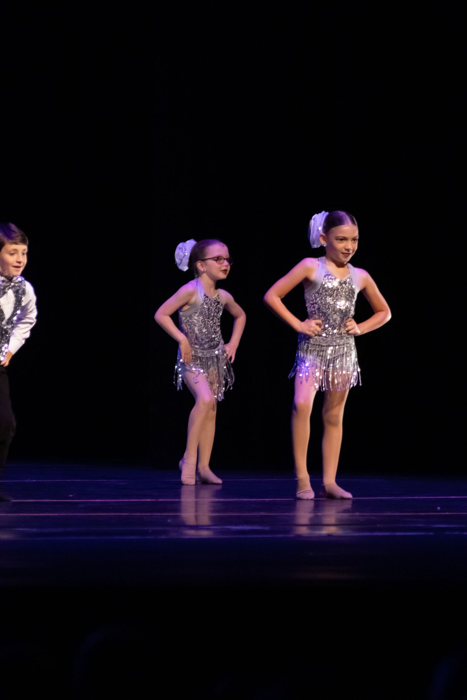 Three young dancers in silver sequin costumes perform on a dark stage during a recital.