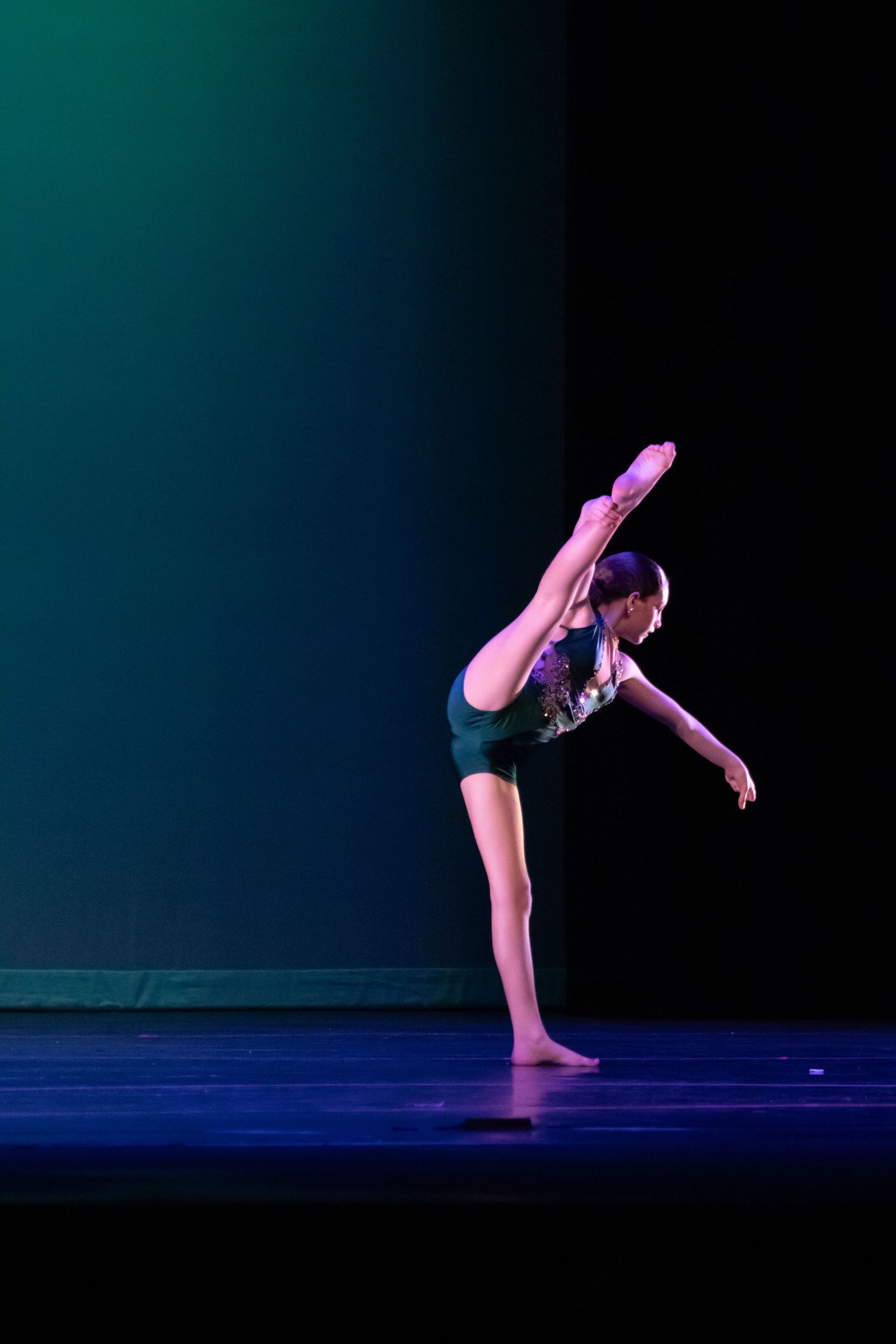A young dancer performs an arabesque in a dark green leotard under teal and blue stage lights.