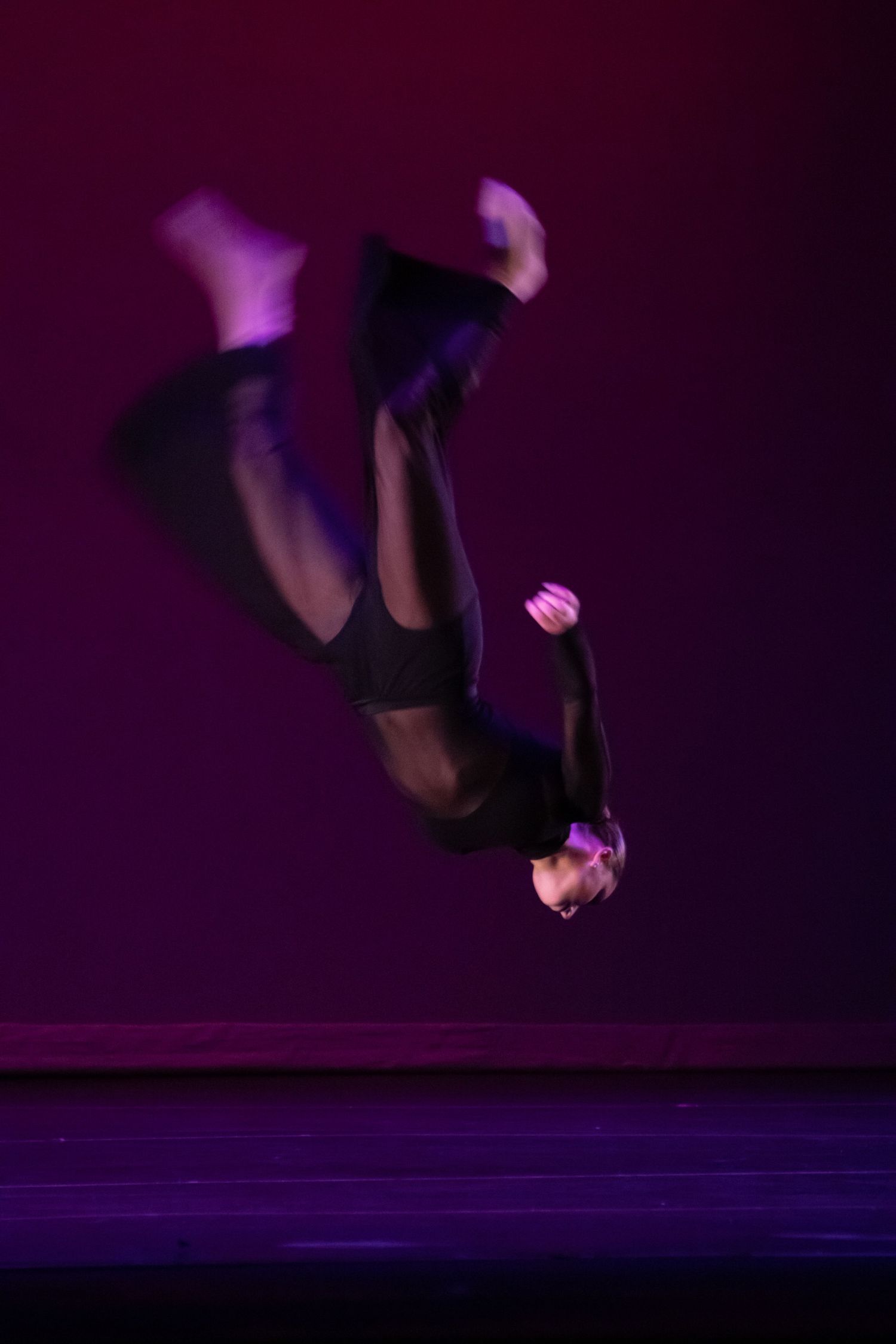 A dancer in a black outfit captured mid-backflip against a deep purple stage background.
