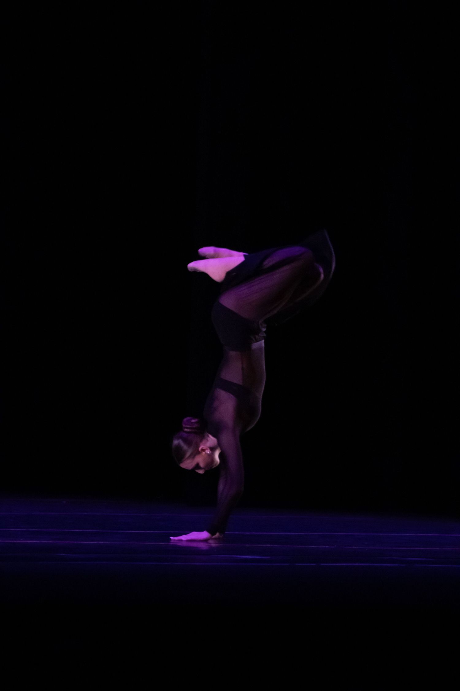 A dancer performs an impressive handstand in all-black attire on a dark stage.