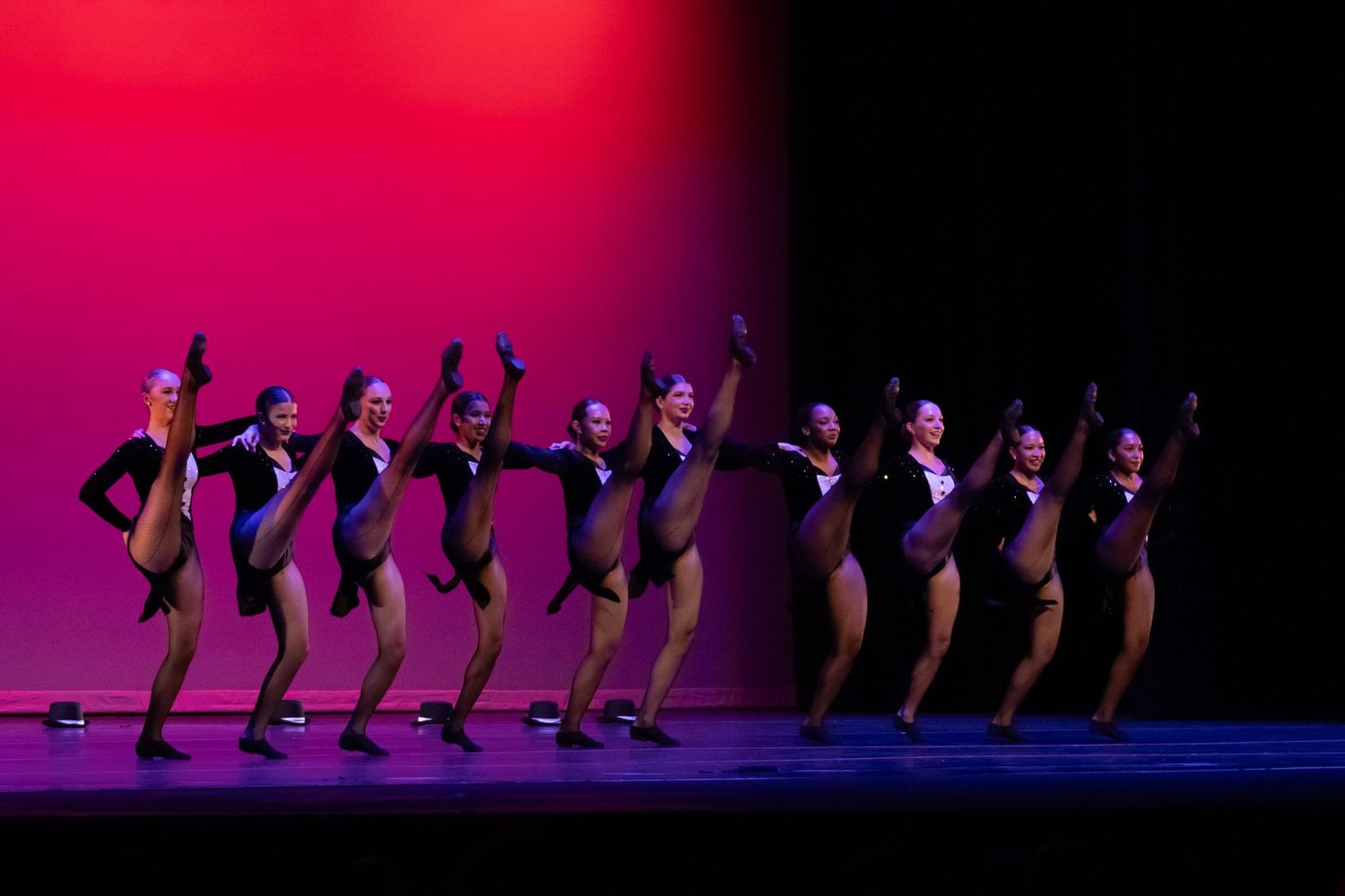 A line of dancers in black costumes perform in unison under red and pink stage lights.