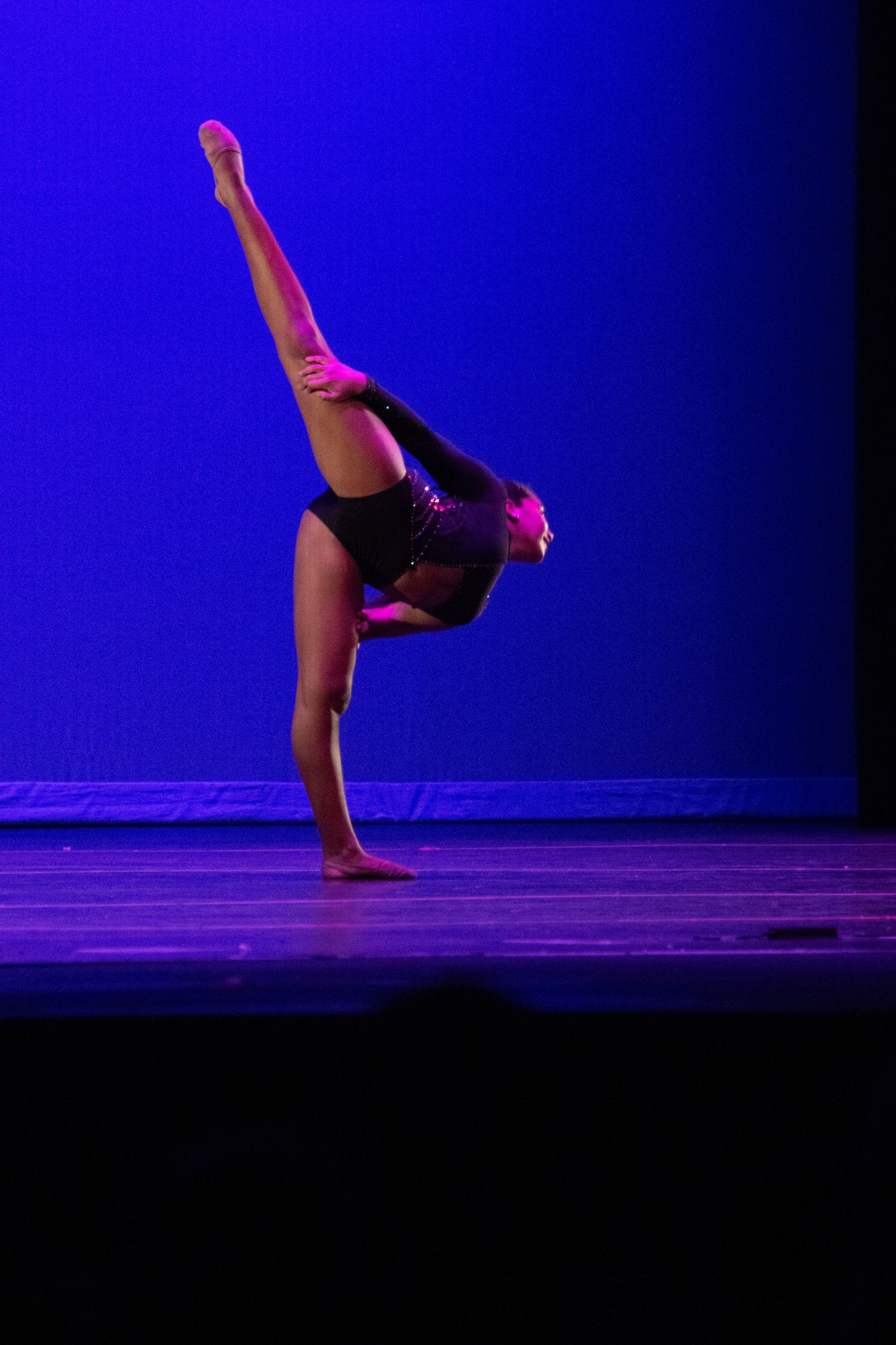 A dancer balances on one foot in a dramatic backbend pose with leg raised high on a blue-lit stage.