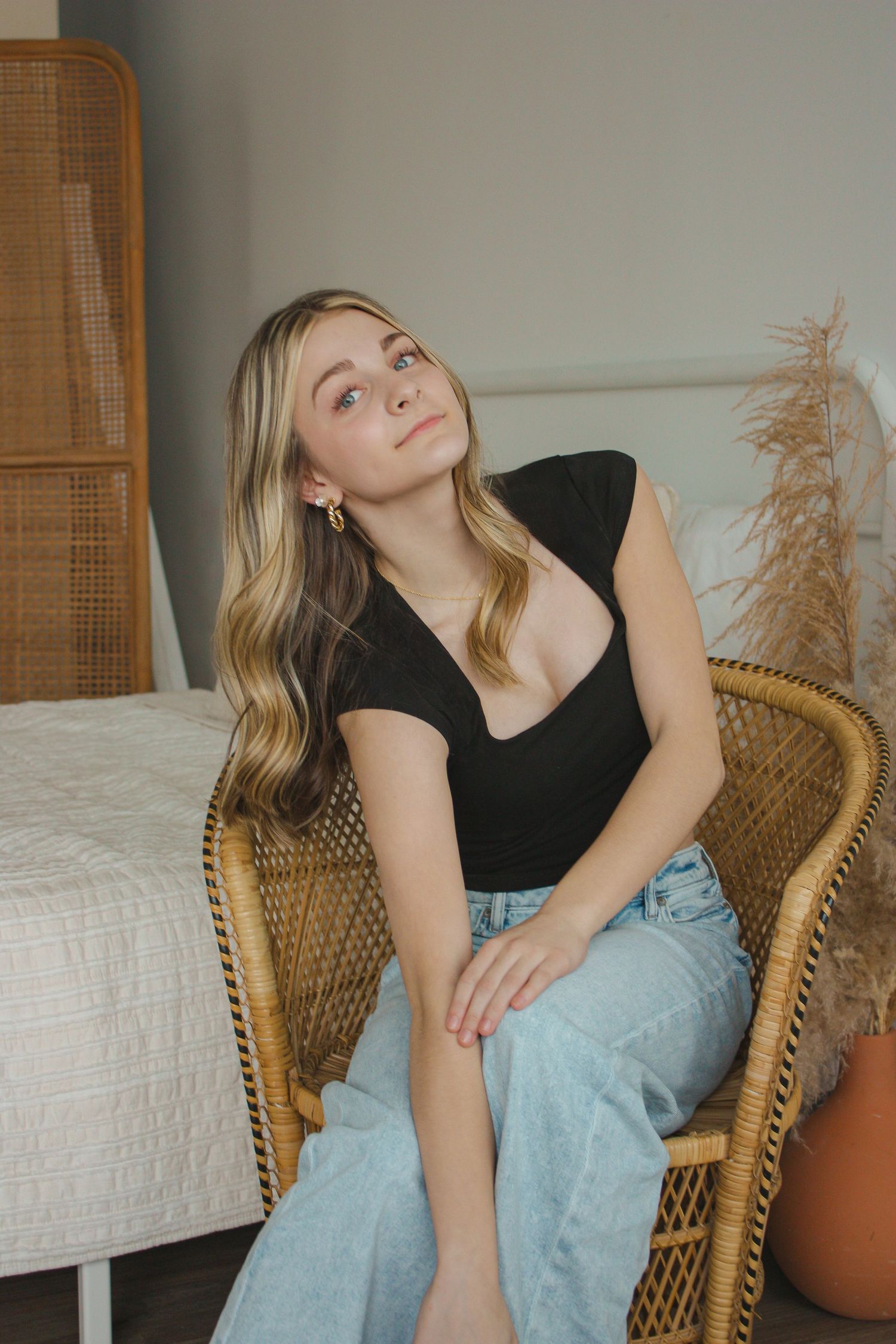 Blonde woman in black top and light jeans sits relaxed in a wicker chair gazing upward in a cozy room.