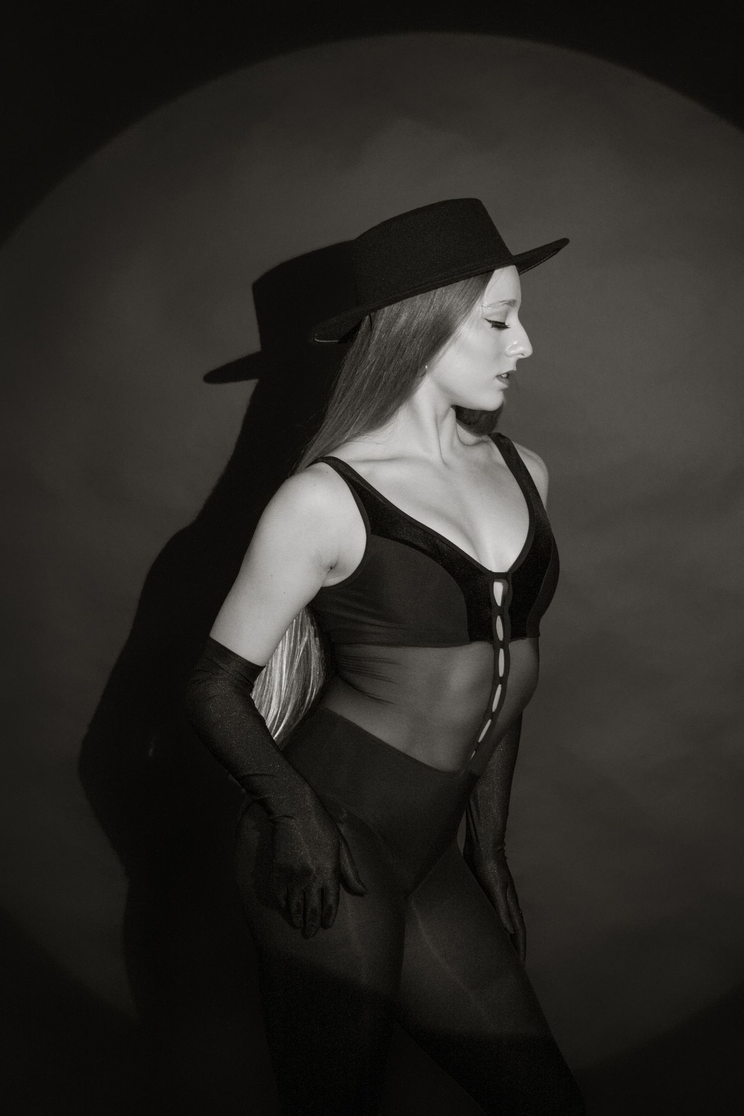 Black and white photo of a woman in lingerie and wide-brim hat posing dramatically against a dark studio background.