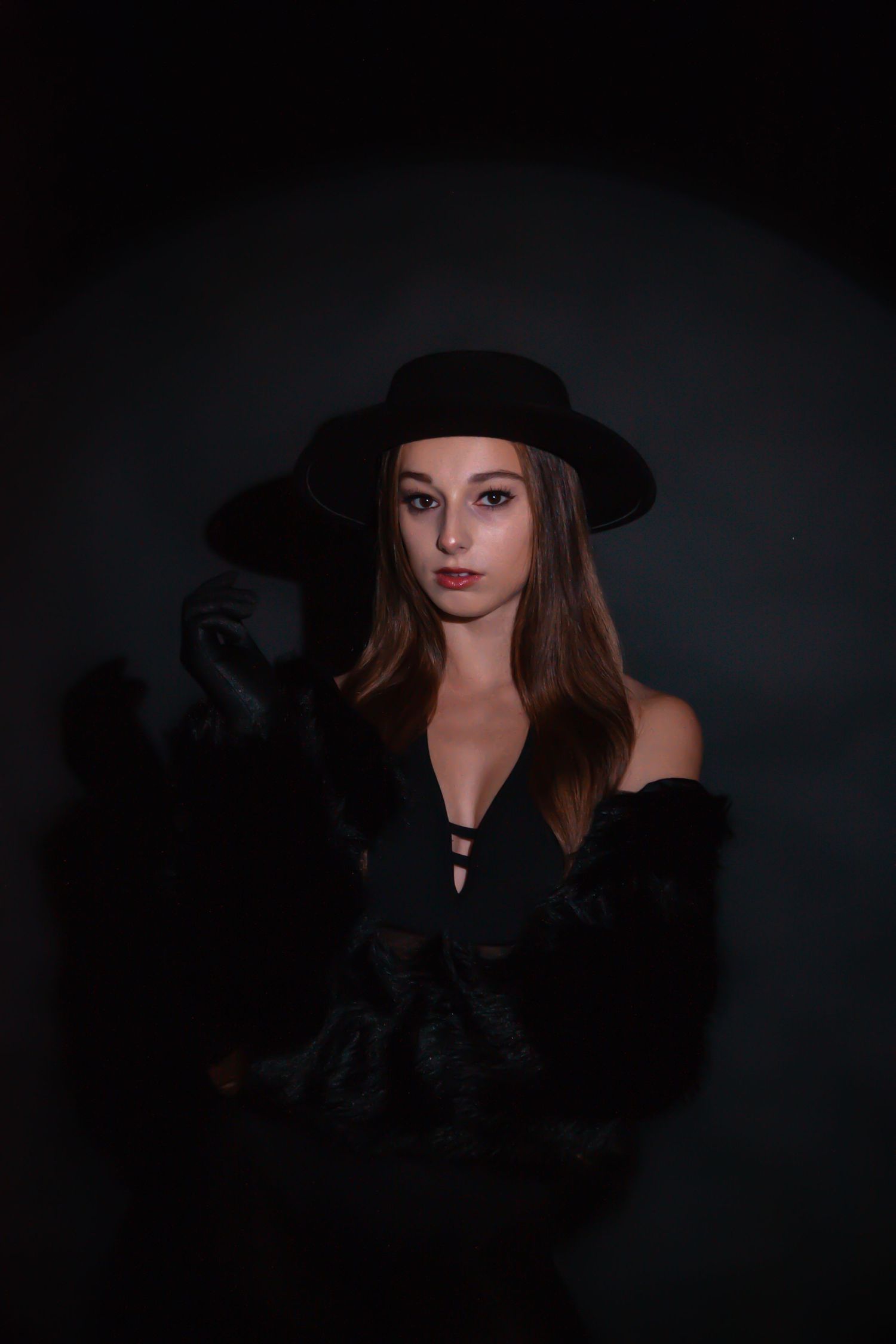 Mysterious woman in black hat and fur coat poses dramatically in dark moody studio setting.