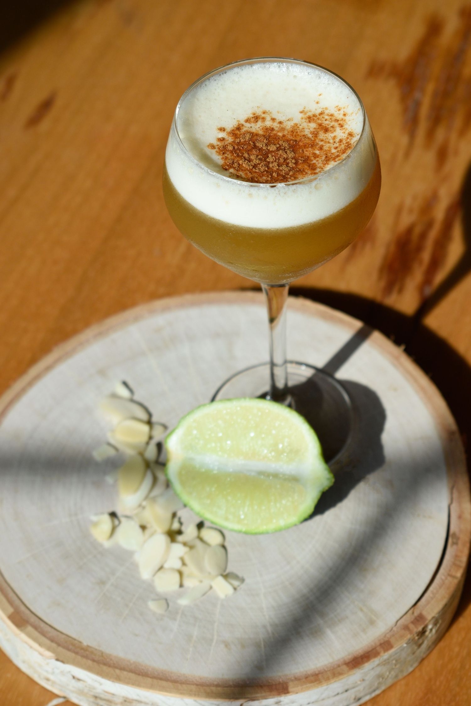 Elegant pisco sour cocktail in a coupe glass topped with cinnamon, served with lime and almonds on a wooden board.