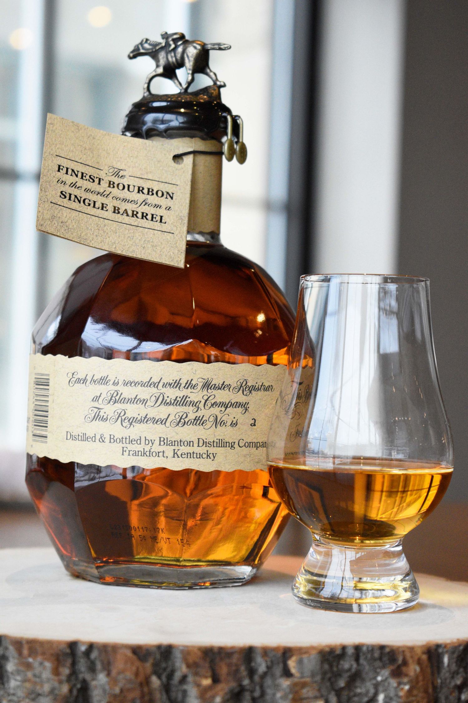 Blanton's Single Barrel Bourbon bottle with neck tag beside a Glencairn glass of whiskey on a wood stump.