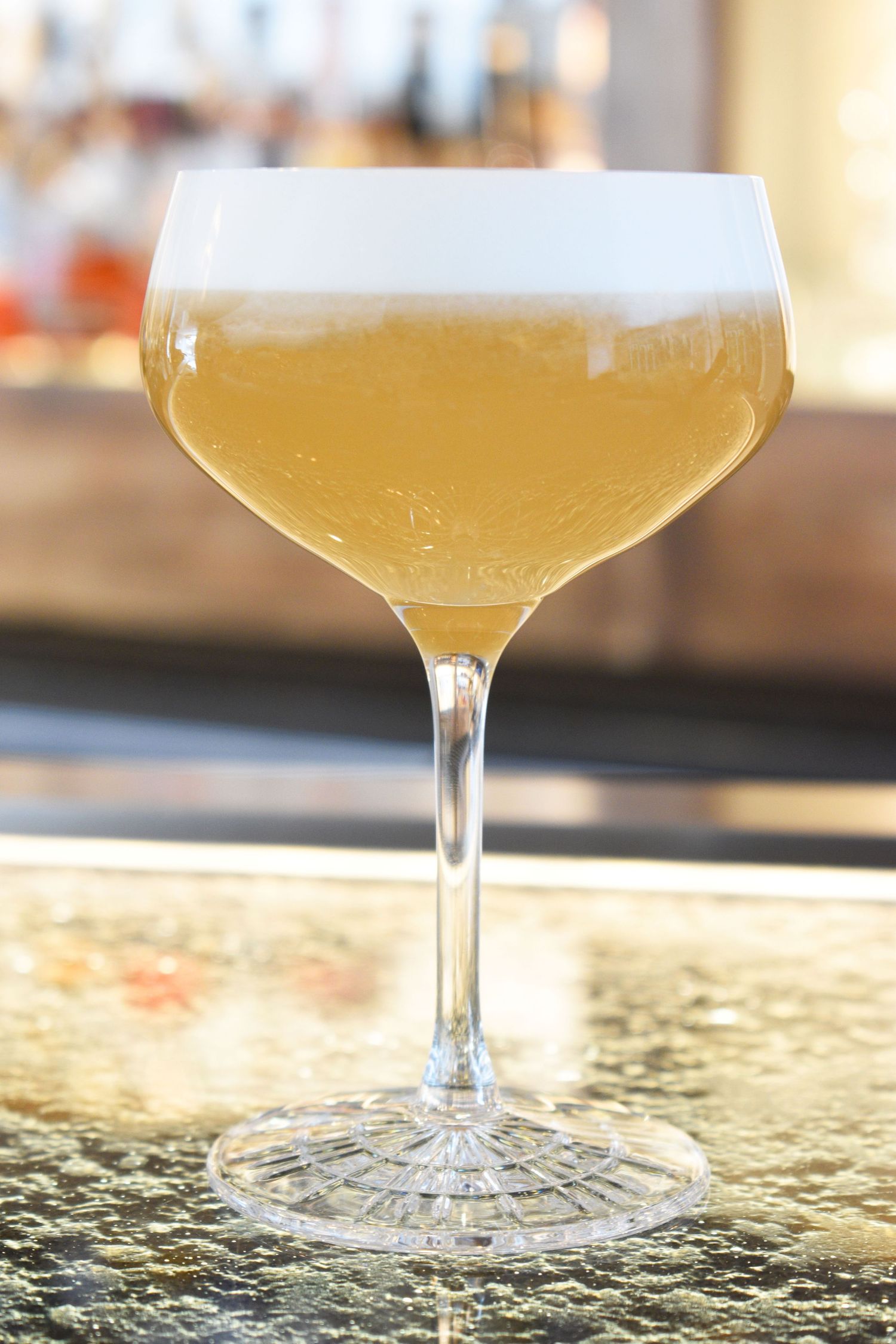 A golden yellow cocktail with frothy white foam served in an elegant crystal coupe glass at a bar.