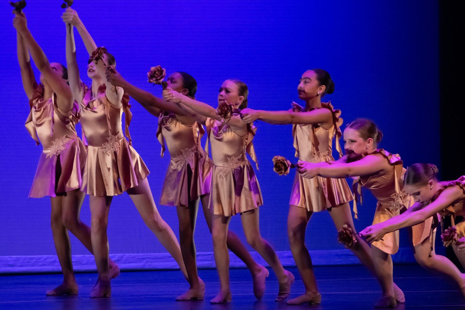 A group of dancers in gold costumes perform a dynamic ensemble piece under blue and pink stage lights.