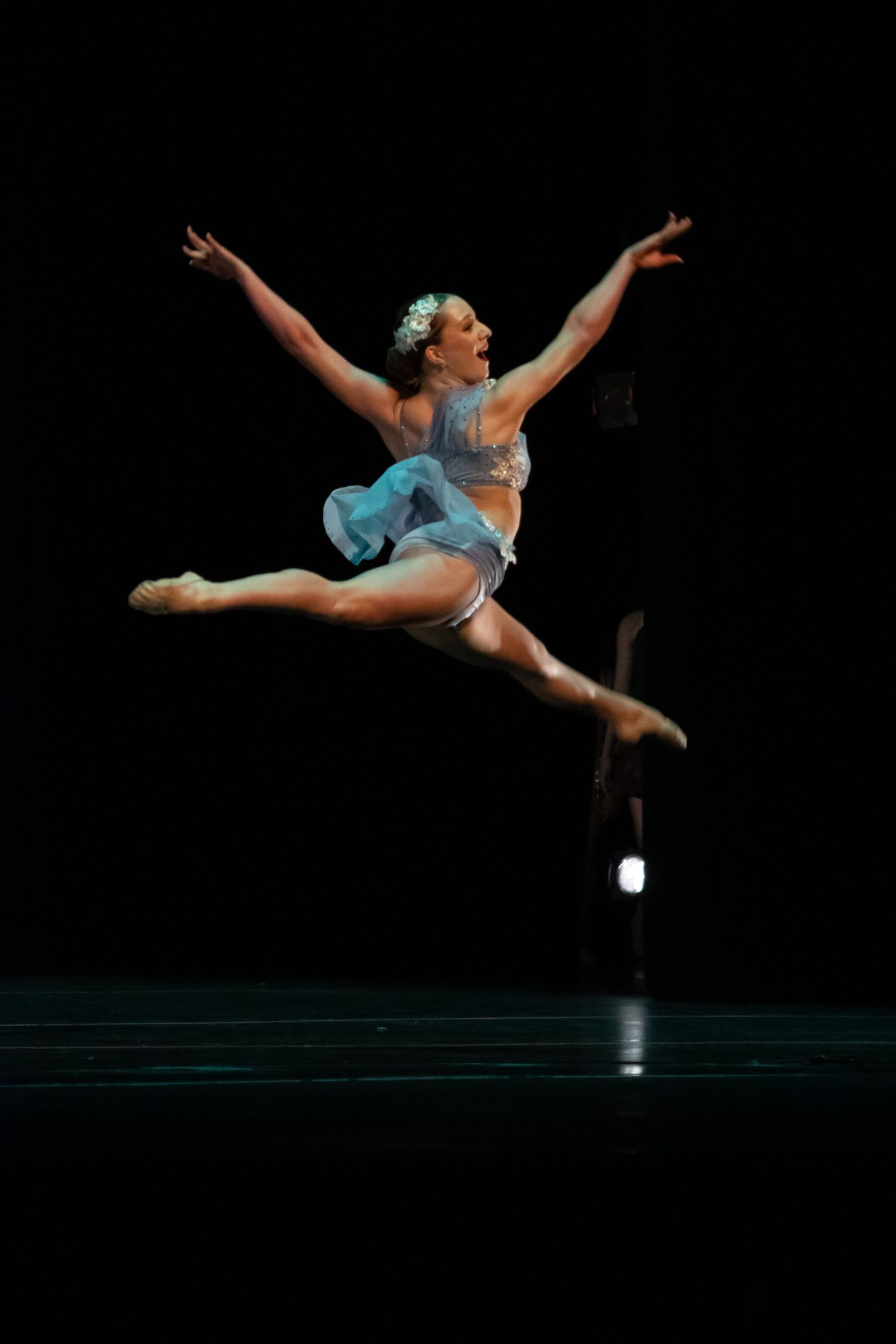 A ballet dancer leaps gracefully in a blue tutu costume against a dramatic black stage backdrop.
