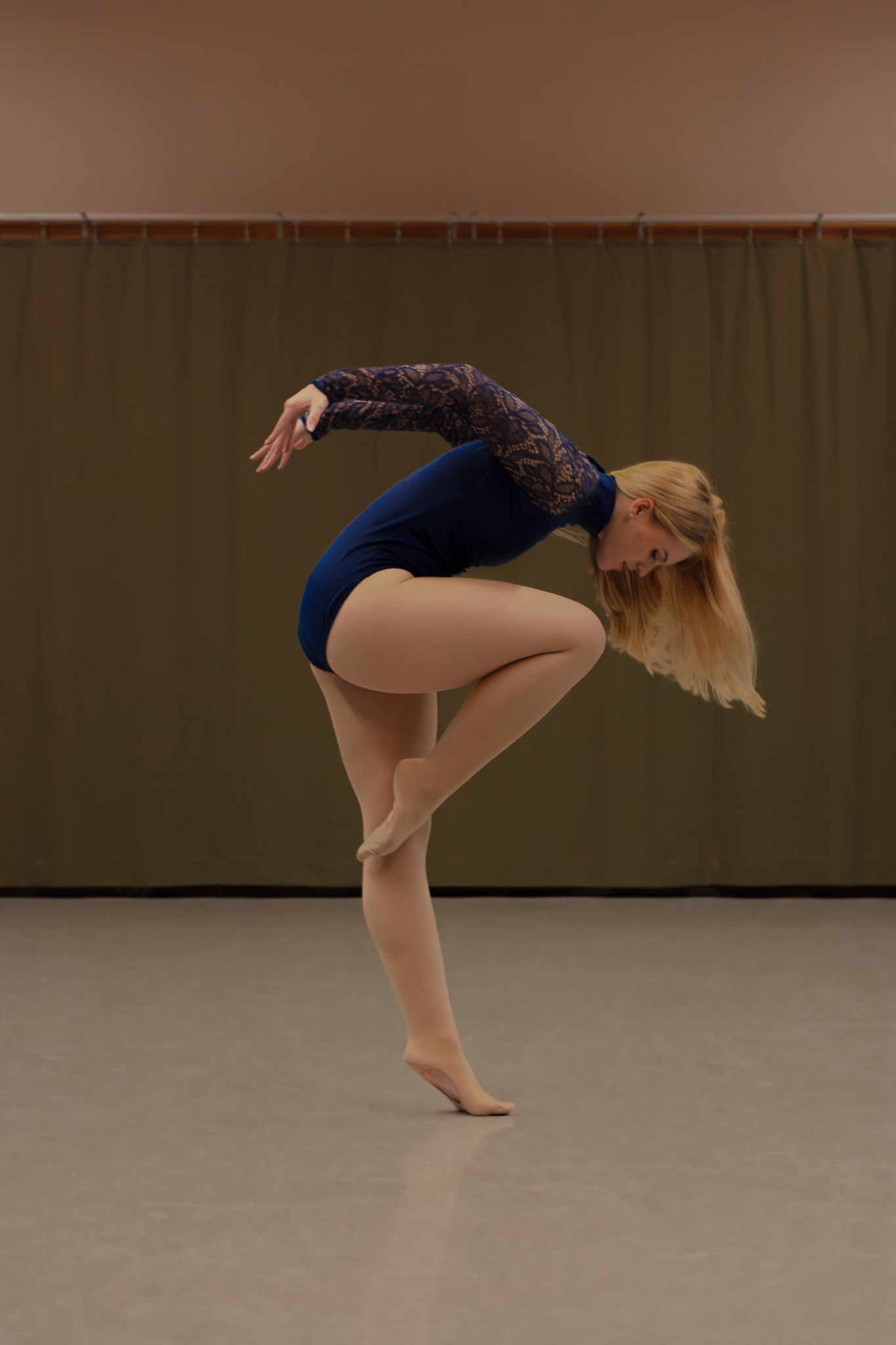 Blonde dancer in blue leotard performs a graceful one-legged balance pose in a dance studio.