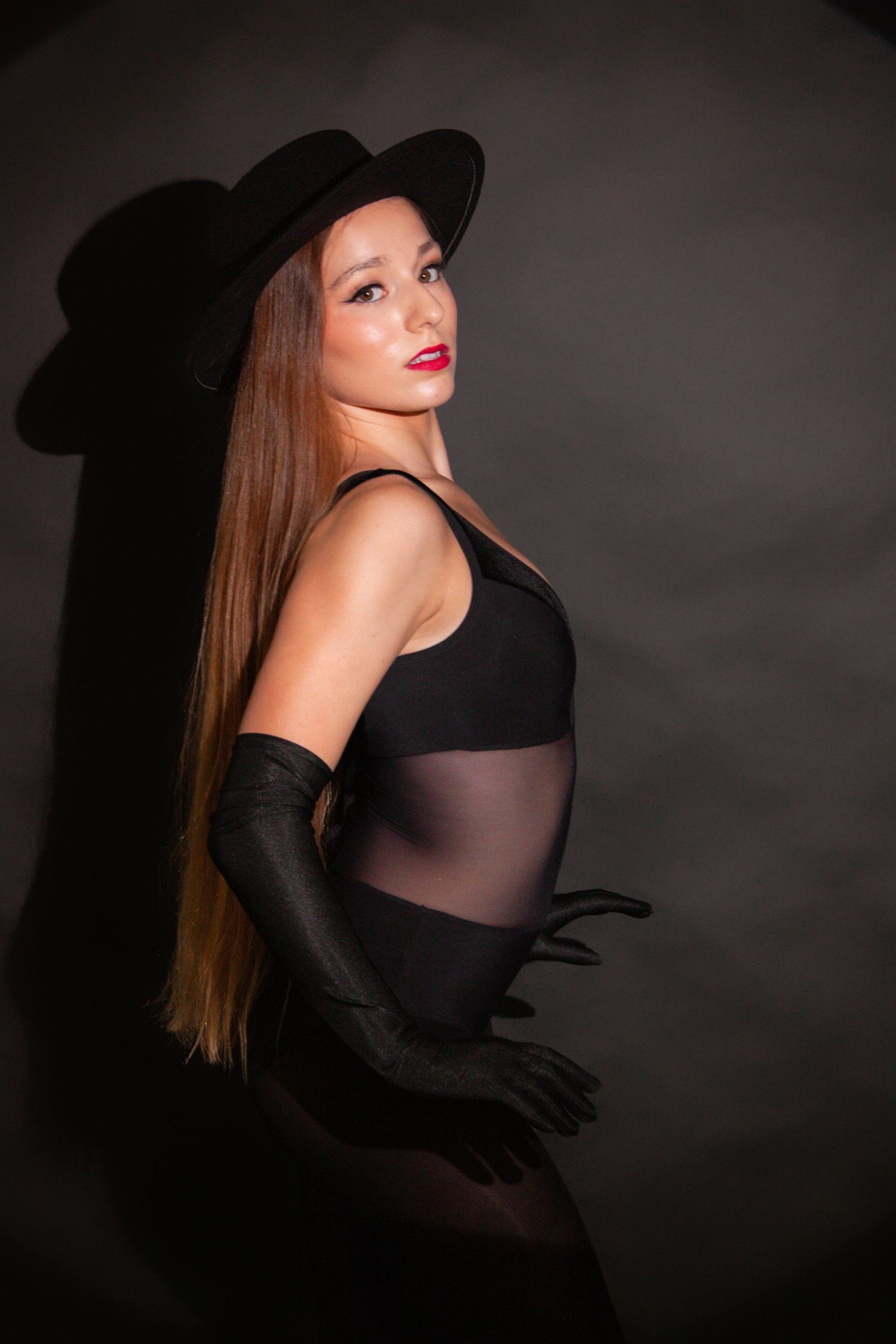 Young woman in black sheer bodysuit and wide-brim hat poses elegantly in a dark studio environment.