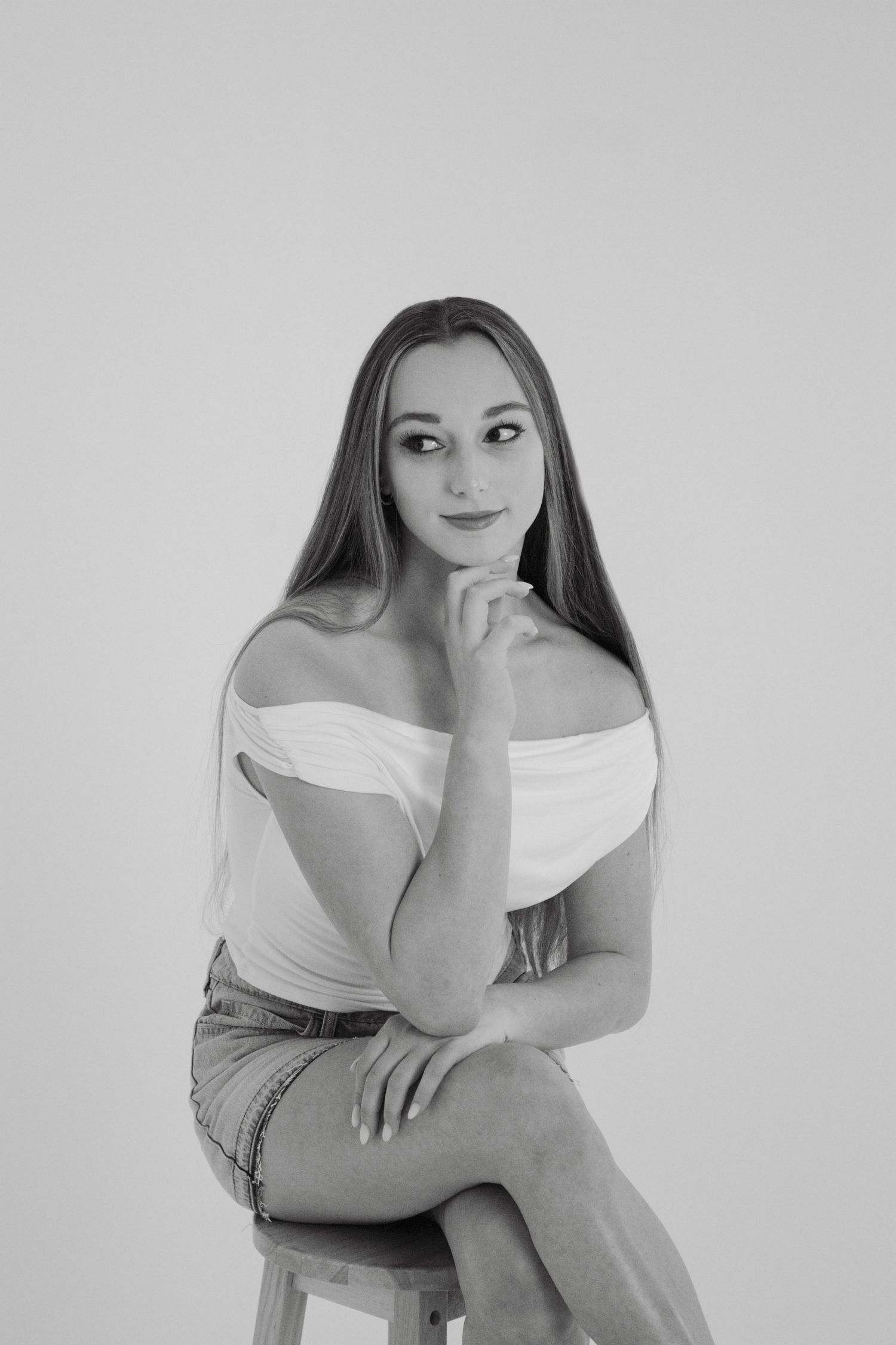 Young woman in white off-shoulder top and denim skirt sits on stool in black and white studio portrait.