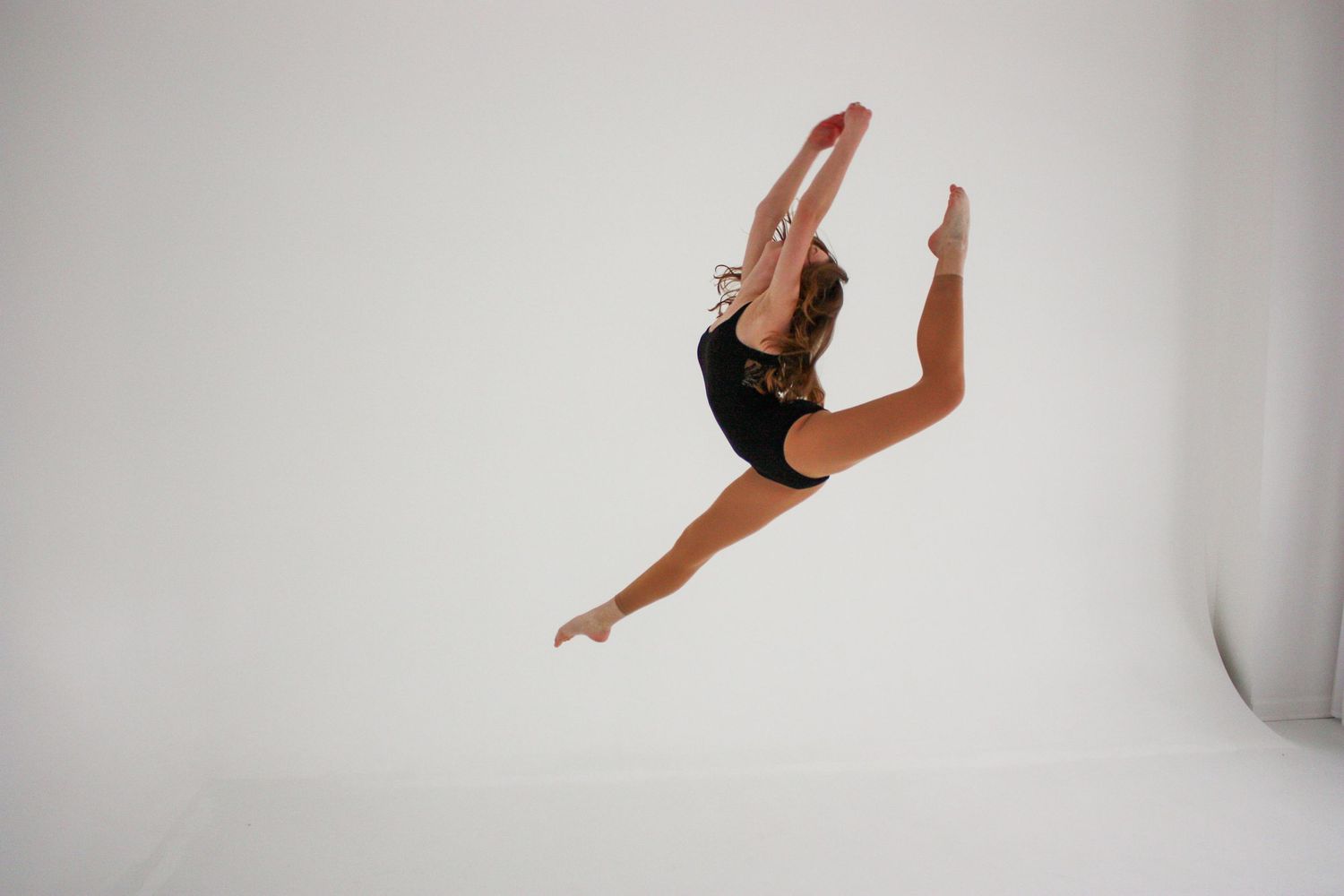 A dancer in a black leotard performs a stunning mid-air split leap against a bright white studio background.