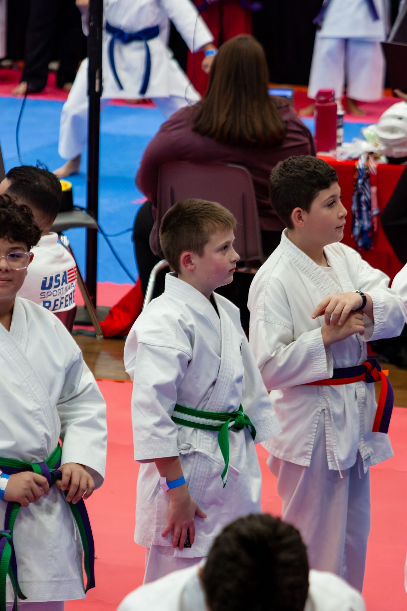 Young karate students in white gi uniforms with green belts stand at attention during a USA karate tournament competition.