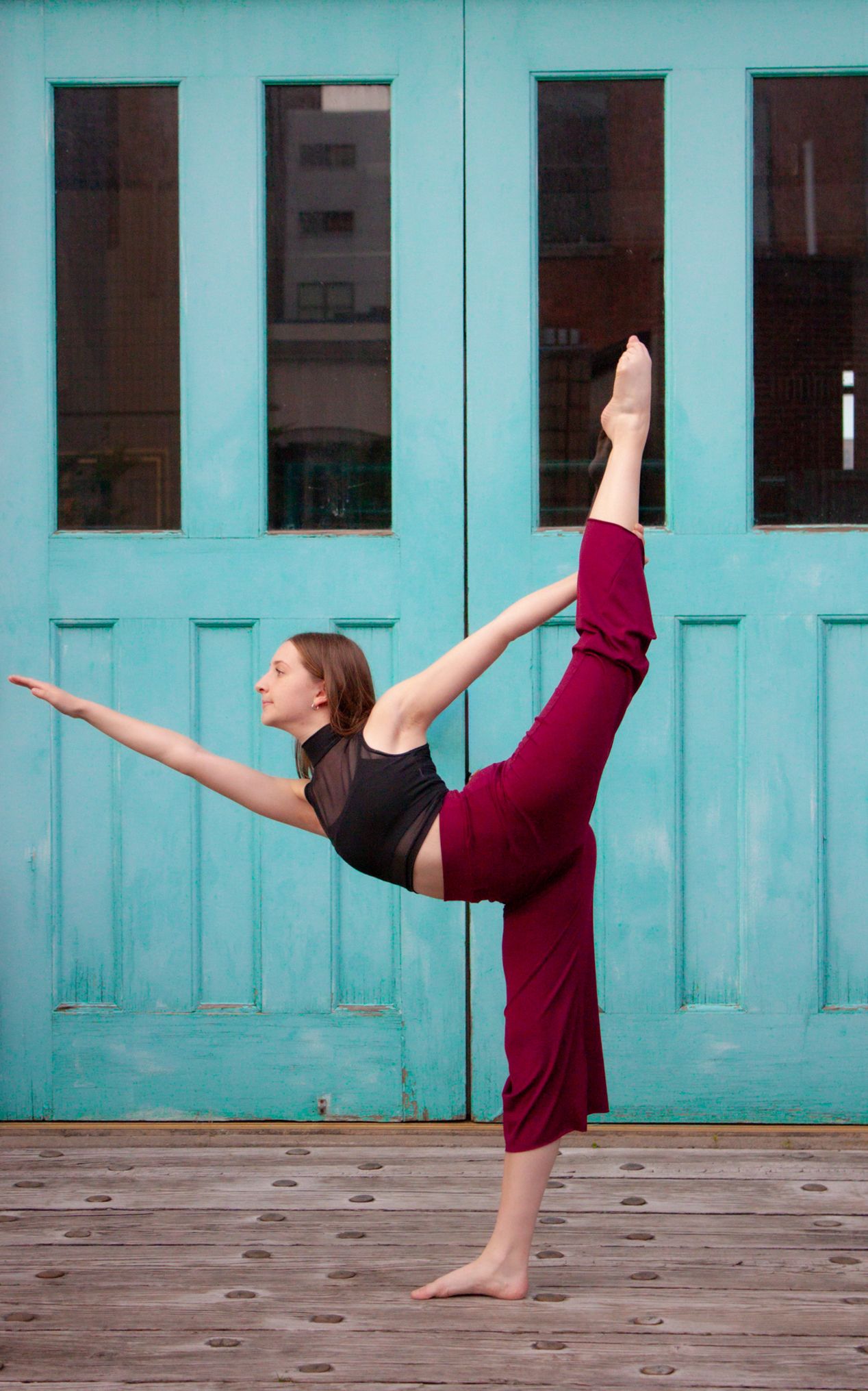 Flexible dancer holds a high kick balance pose in front of turquoise double doors on a wooden deck.