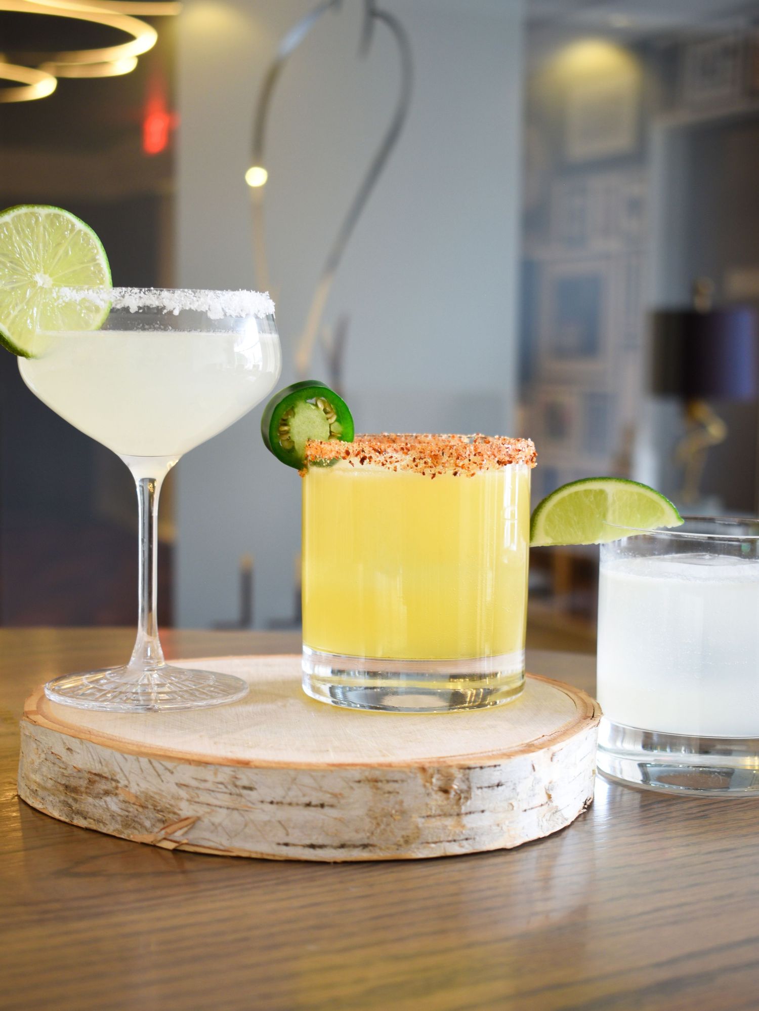 Three cocktails on a wood slice including a yellow margarita with chili rim and jalapeno garnish.