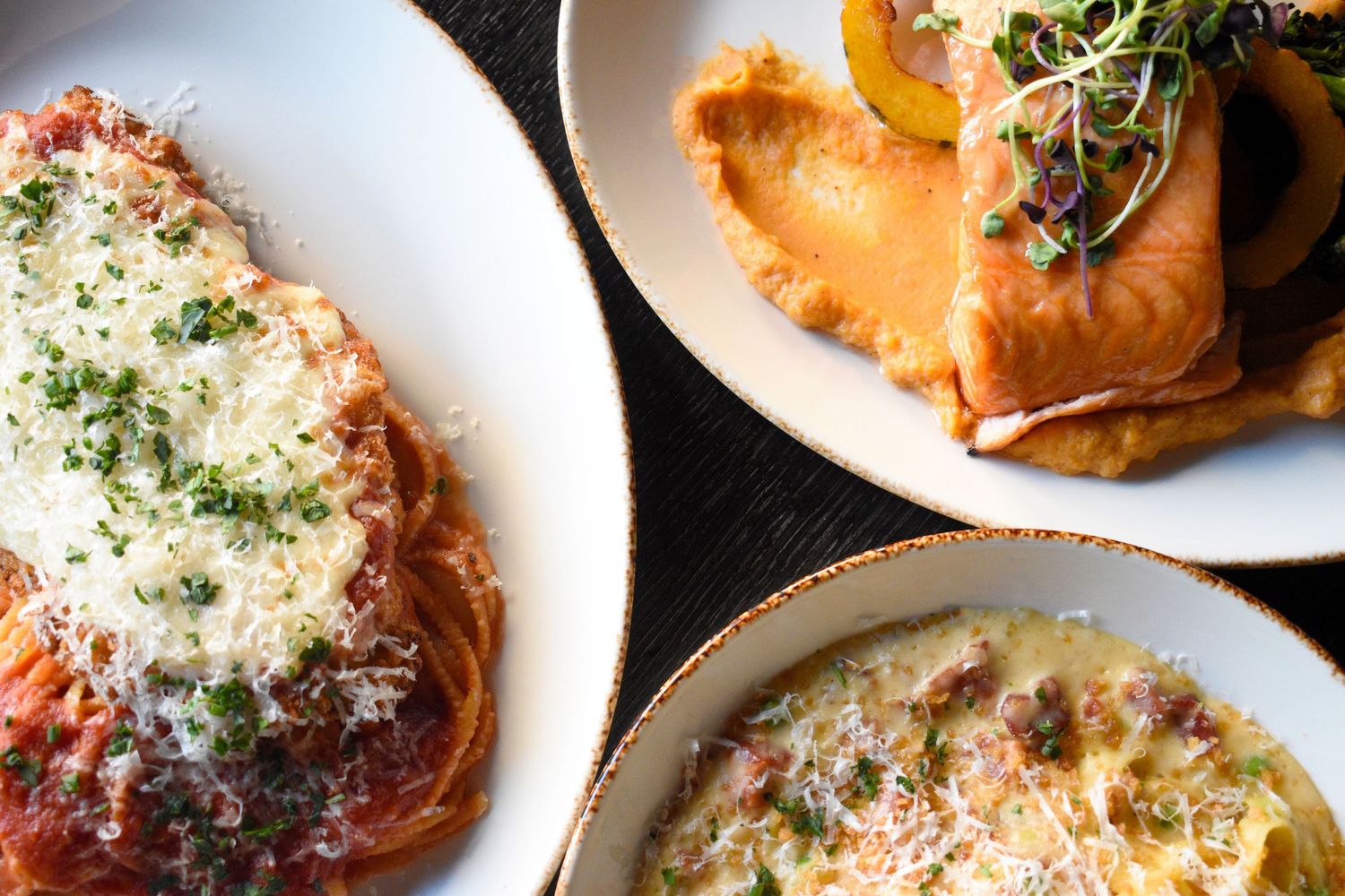 Three Italian dishes including chicken parmesan, pasta, and a creamy entree on a dark table.