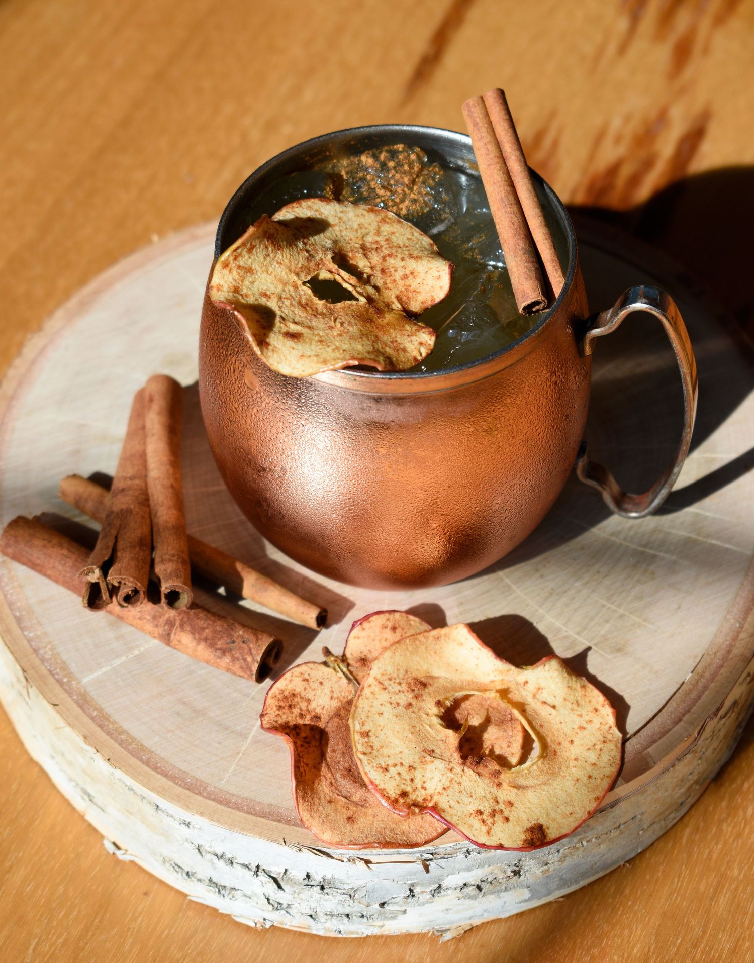 Apple cider Moscow mule in a copper mug garnished with dried apple chips and cinnamon sticks on a wood slice.