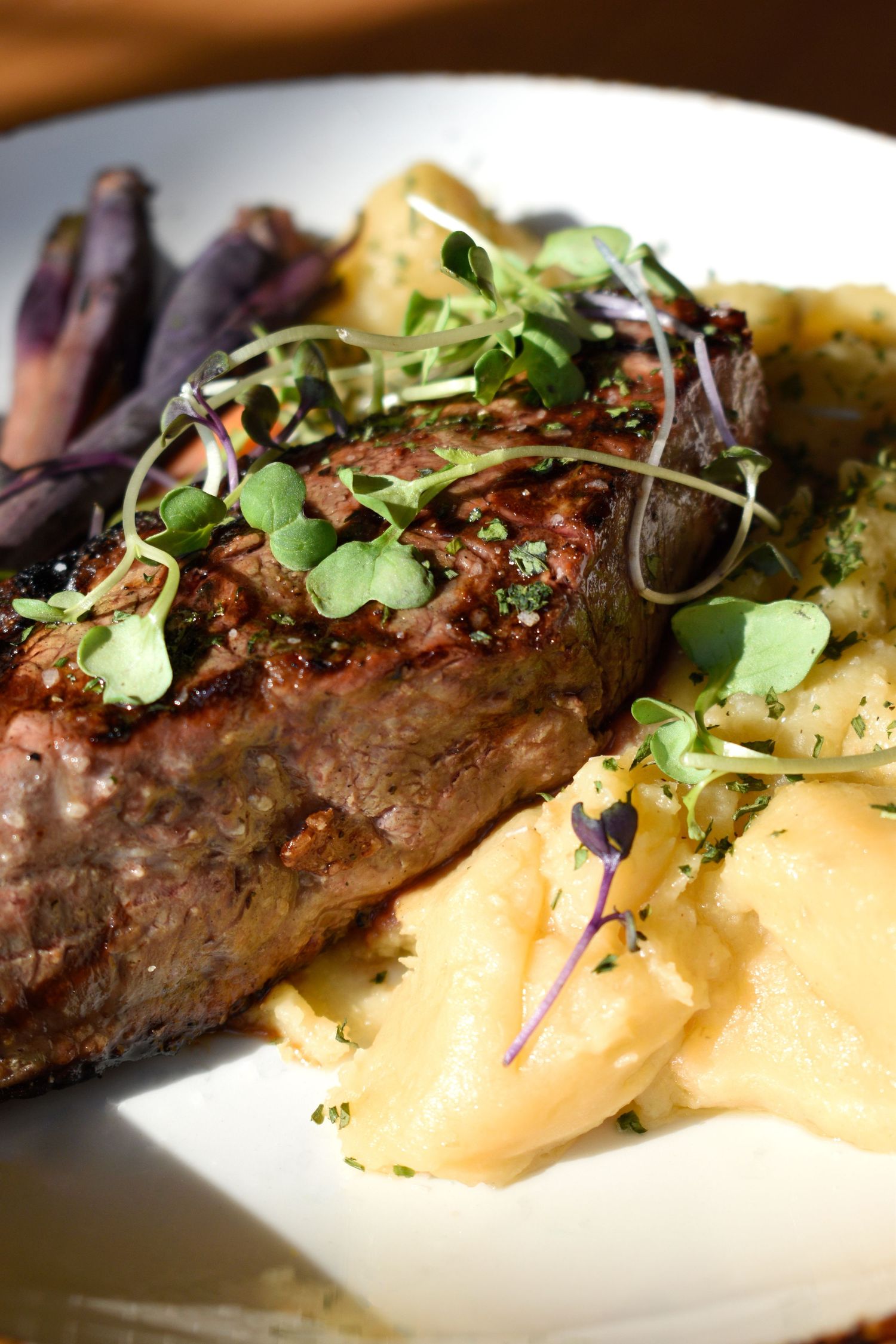 A grilled steak served with creamy mashed potatoes, microgreens, and roasted purple vegetables.
