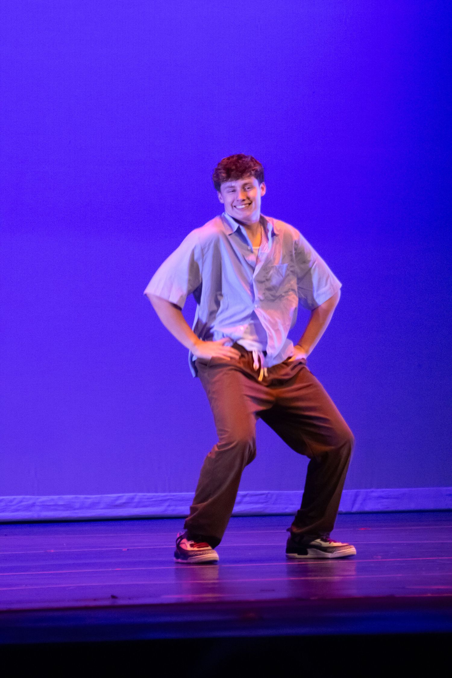 A smiling male dancer poses confidently in brown pants and blue shirt on a purple-lit stage.