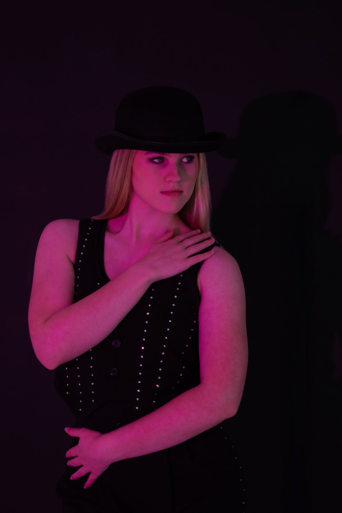 Blonde woman in black studded vest and bowler hat under pink neon light.