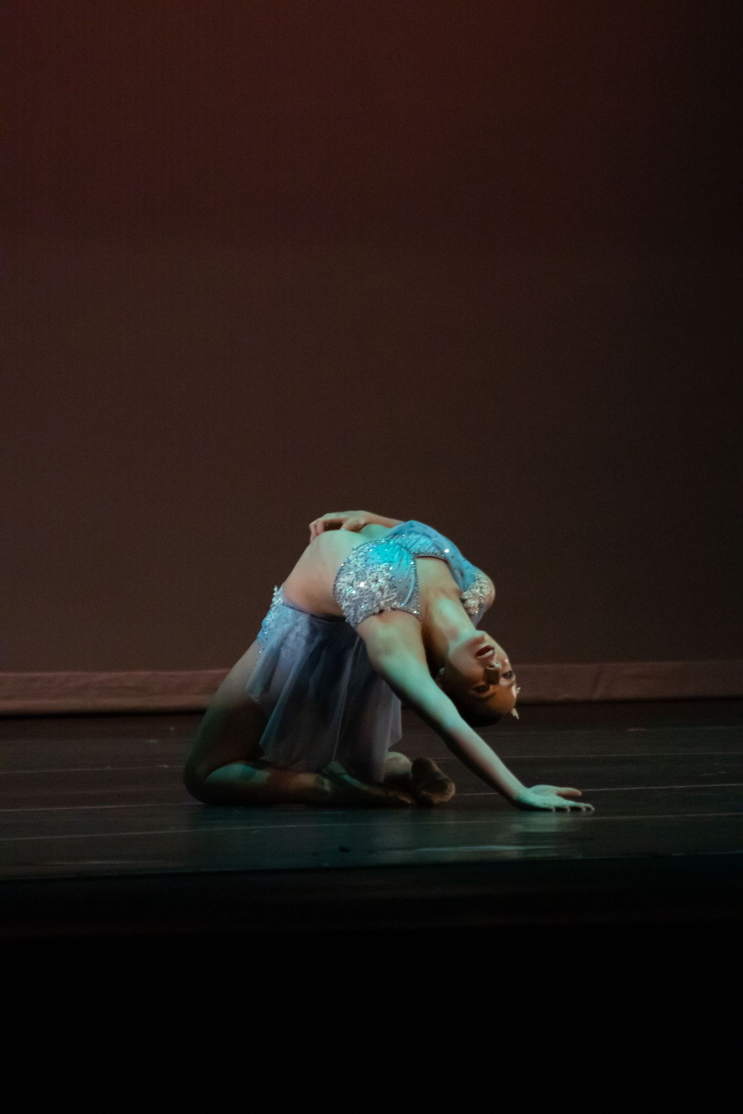 Flexible dancer in blue sequined costume performs a deep floor contortion pose.
