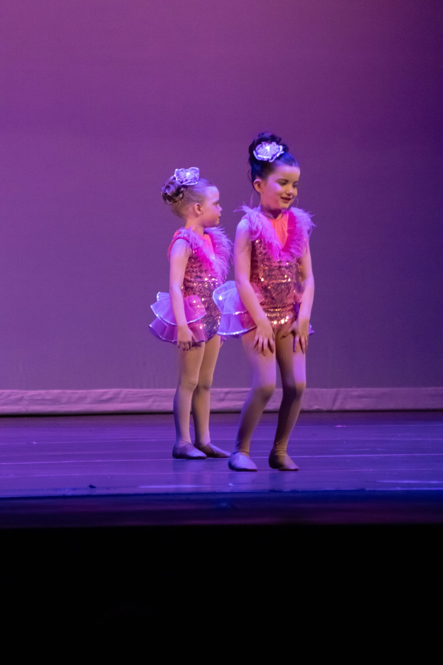 Two young girls in pink sequin dance costumes perform on a colorful stage during a dance recital.