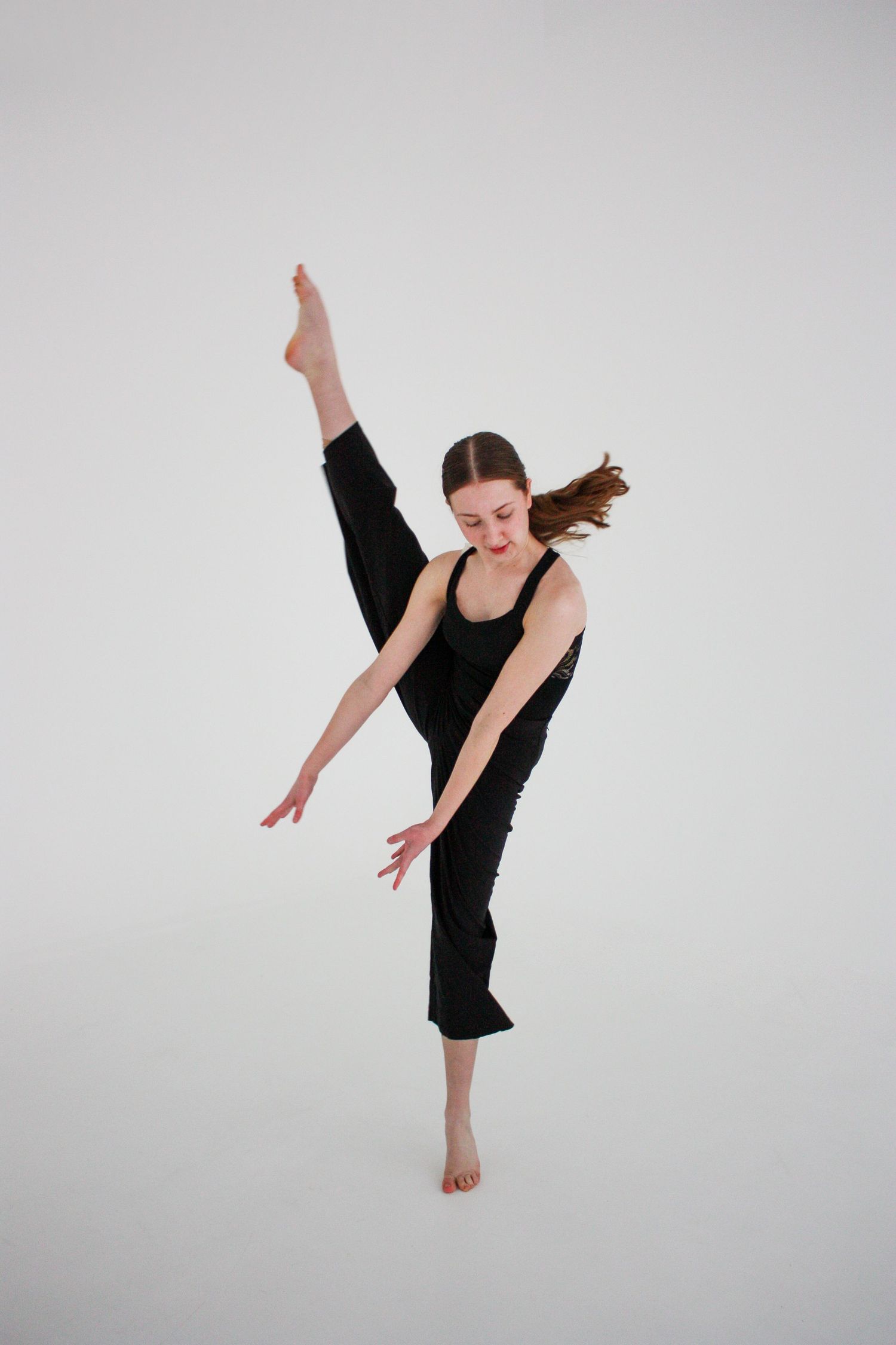 Young dancer in black outfit performs high kick balance on white background.