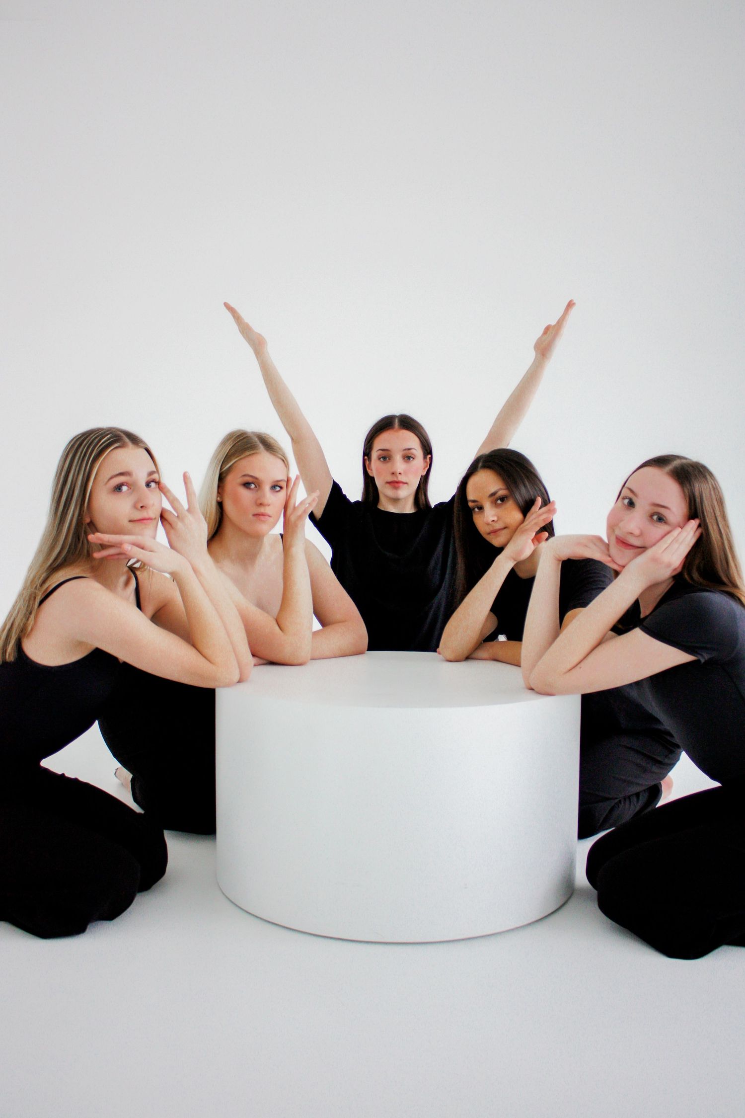 Five female dancers in black outfits pose playfully around a white cylindrical prop in a studio.