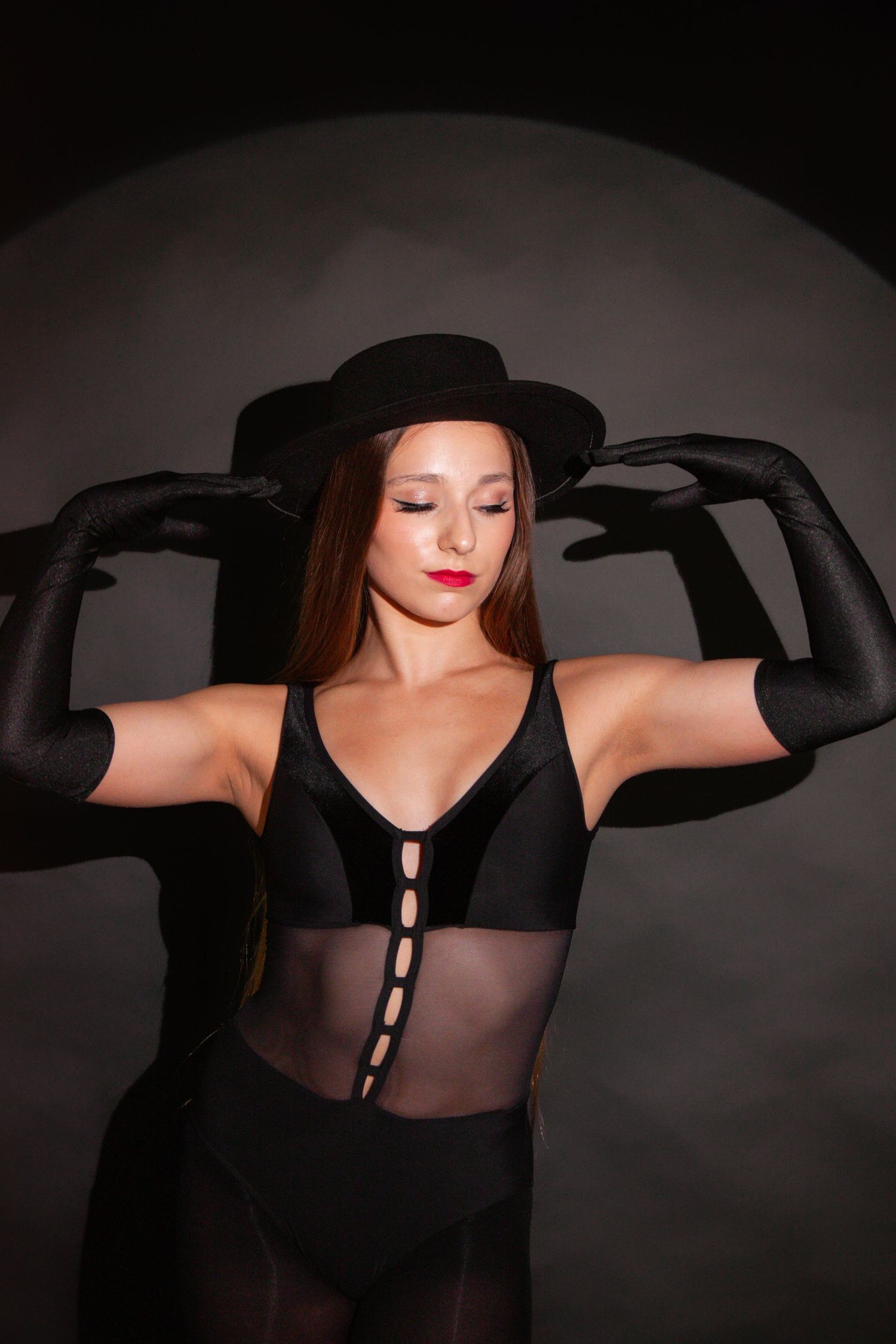 Dancer in black bodysuit and hat poses dramatically with black gloves in a dark studio setting.