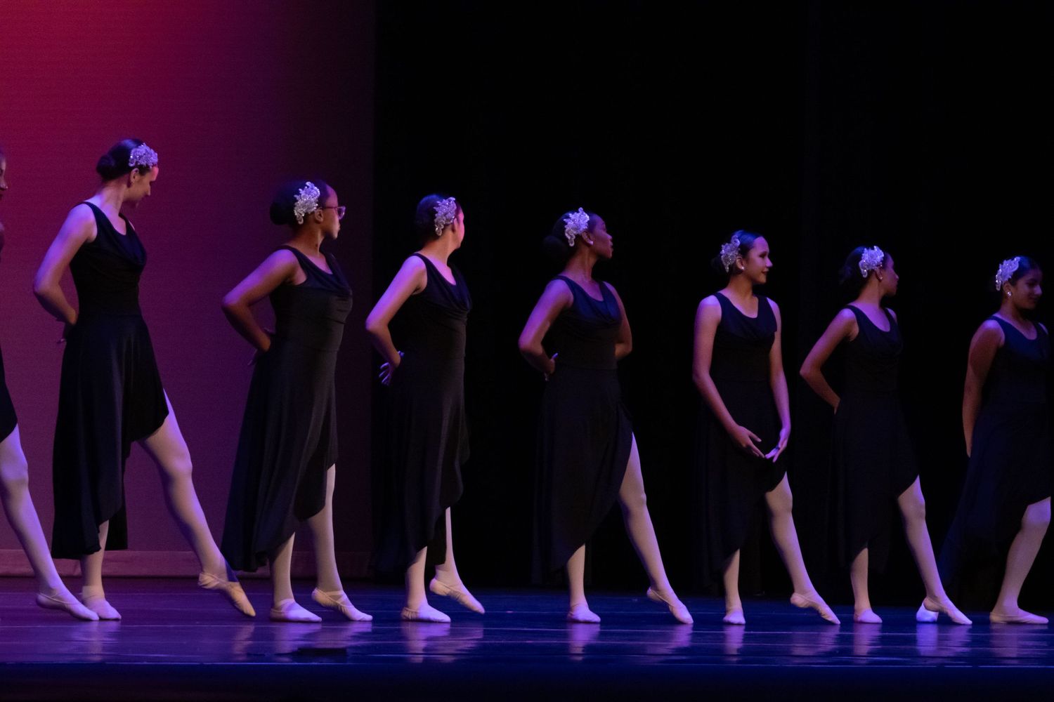 Dancers in black dresses perform on stage in a synchronized line during a dance recital.