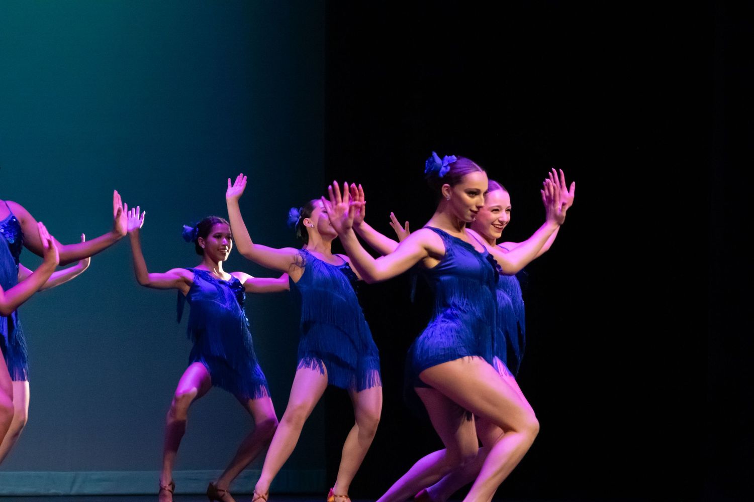Female dancers in blue fringe outfits perform an expressive contemporary routine on a darkened stage.