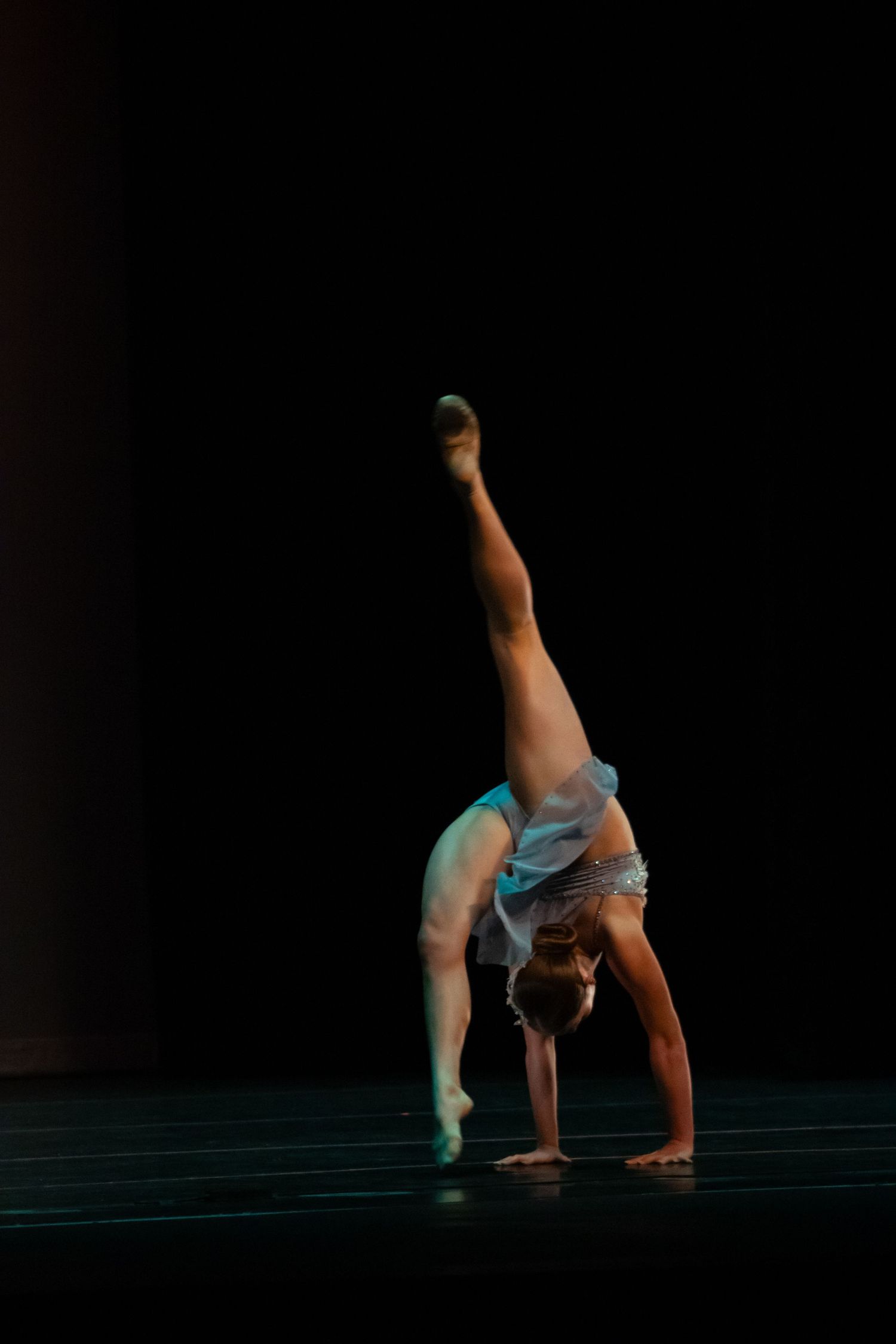 A dancer executes a one-handed handstand with leg extended high under dramatic dark stage lighting.