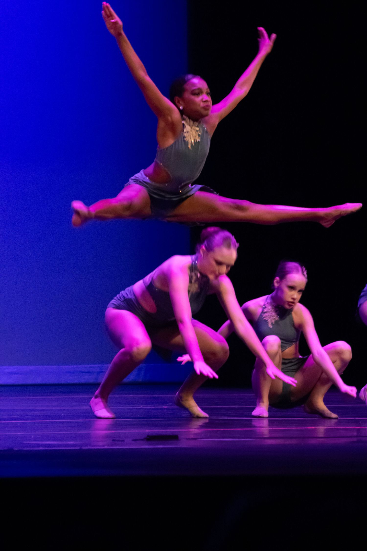 A dancer performs a stunning leap over crouching teammates in gray costumes on a lit stage.