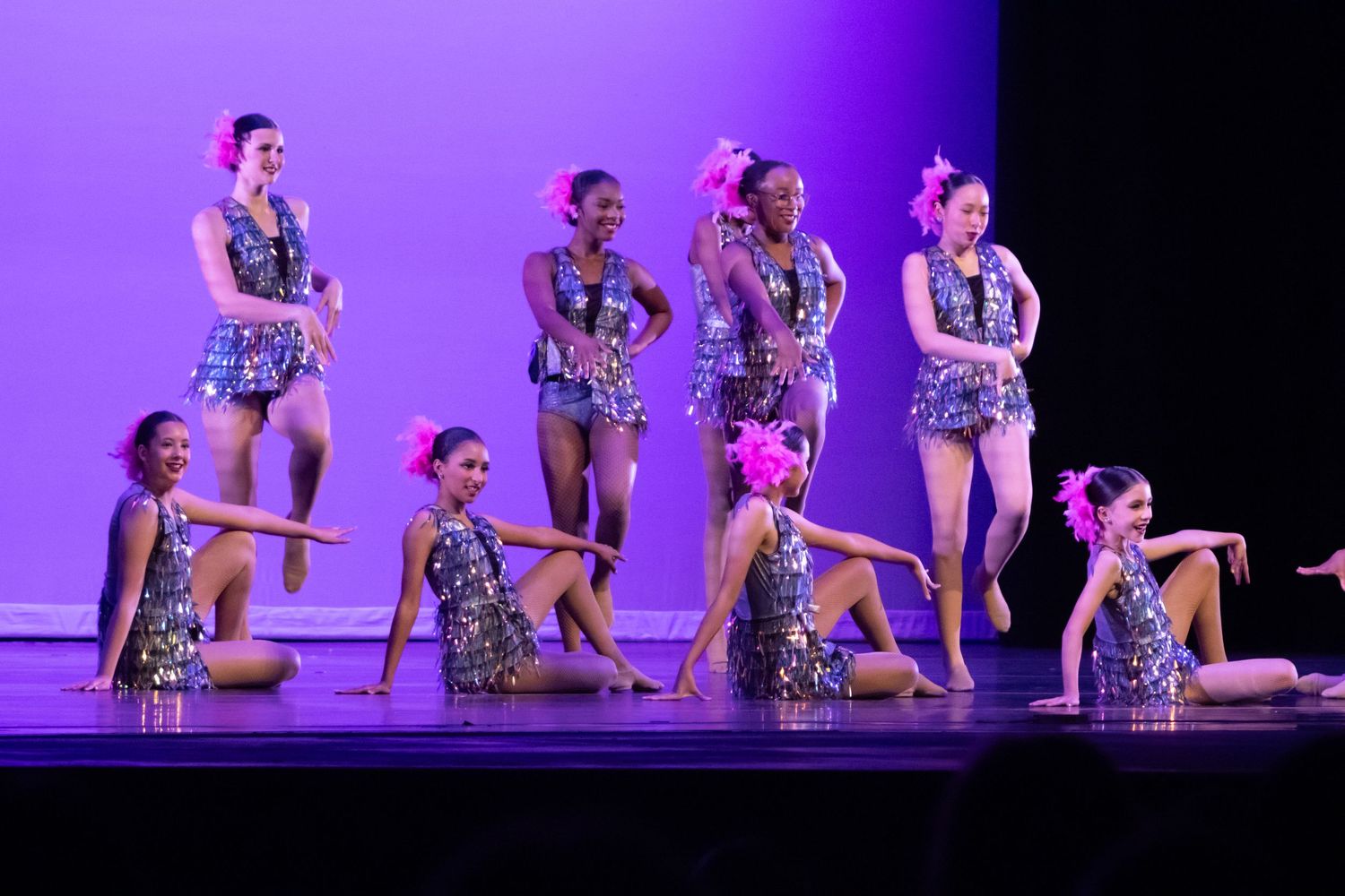 Nine dancers in sparkly silver costumes strike poses on a purple-lit performance stage.