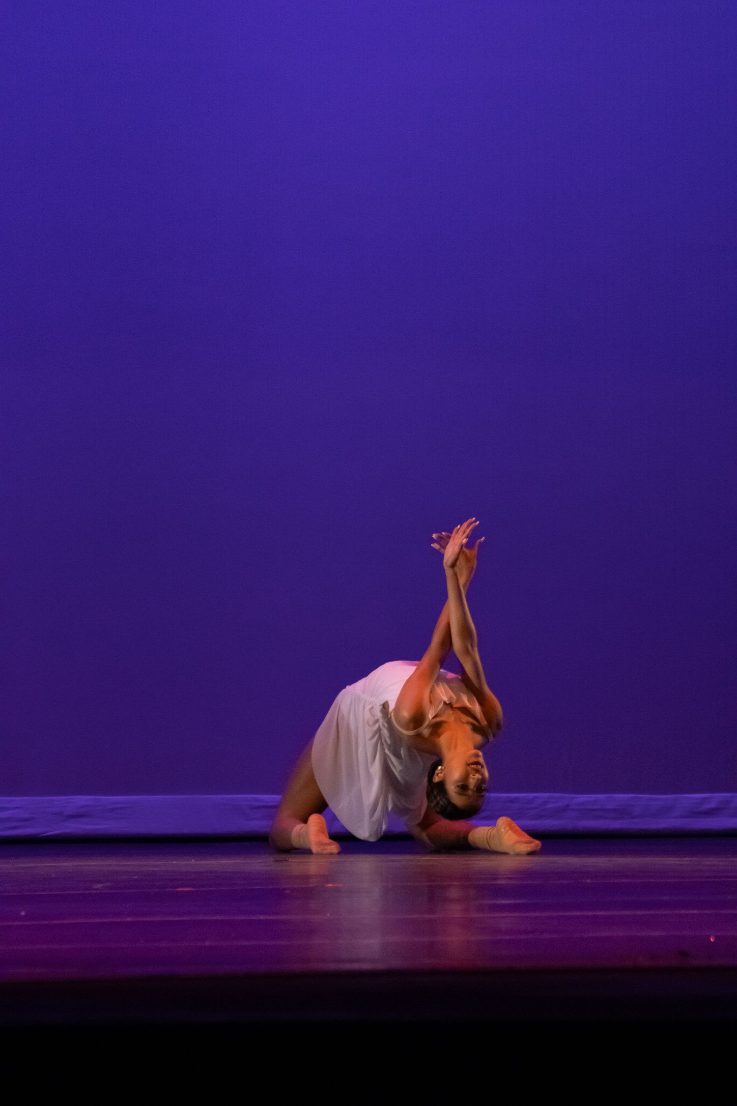 A dancer performs a dramatic floor pose on stage, arching backward with arms raised against a purple backdrop.