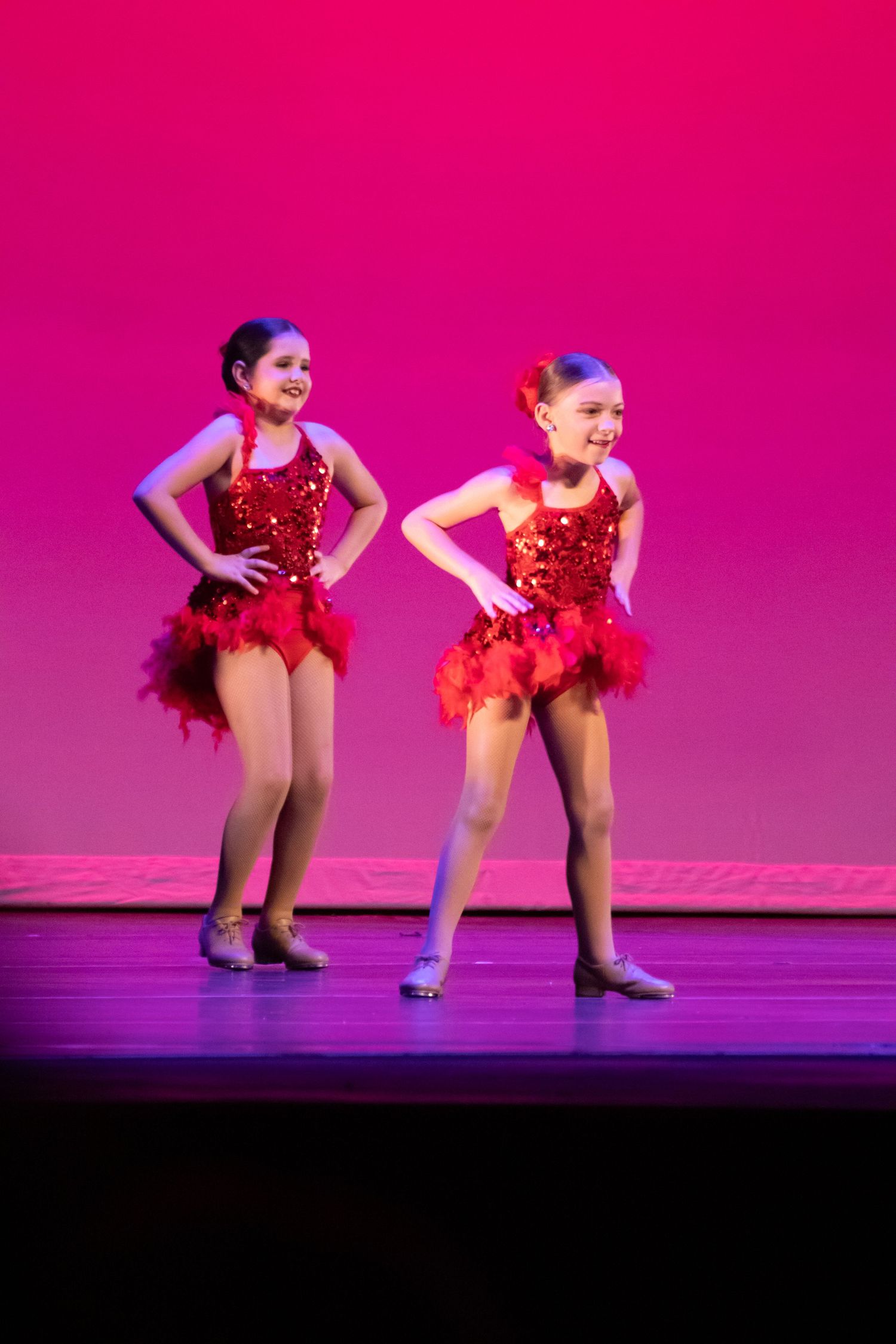 Two young girls smile on stage in matching red sequined tutus during a dance recital performance.
