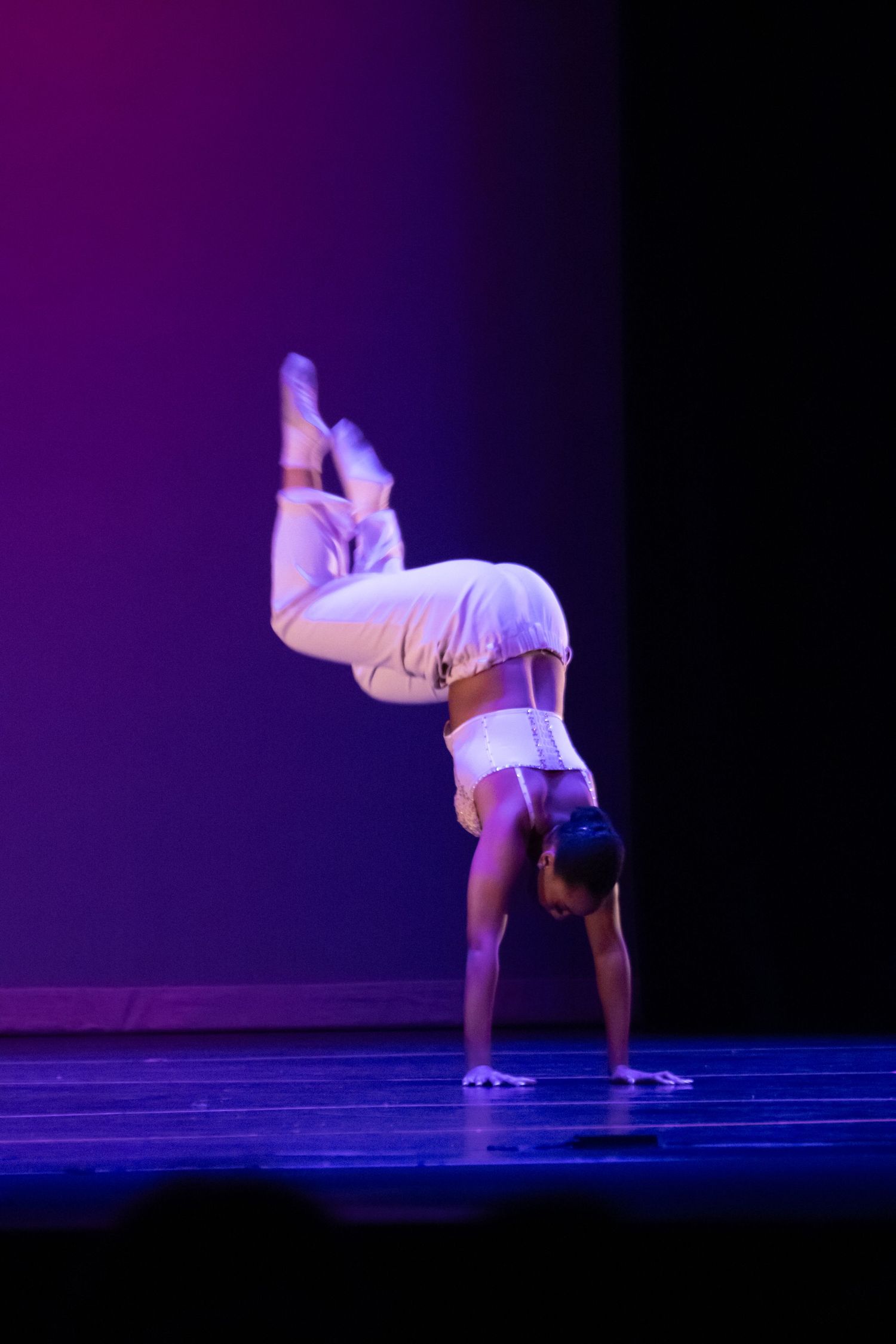 Acrobatic dancer in white outfit executes a handstand with bent knees on purple-lit stage.