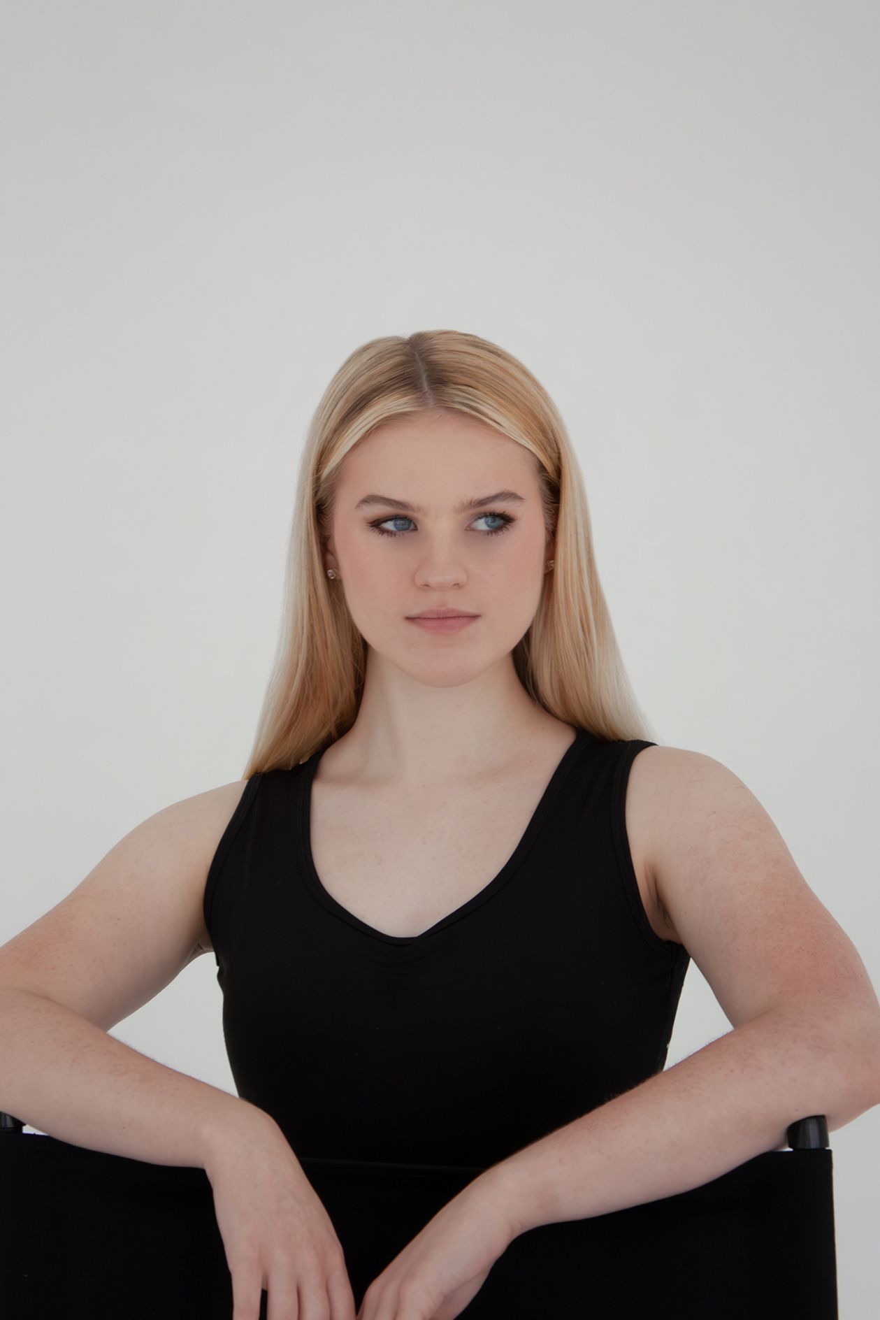 Blonde female dancer seated in black tank top, gazing sideways against white background.