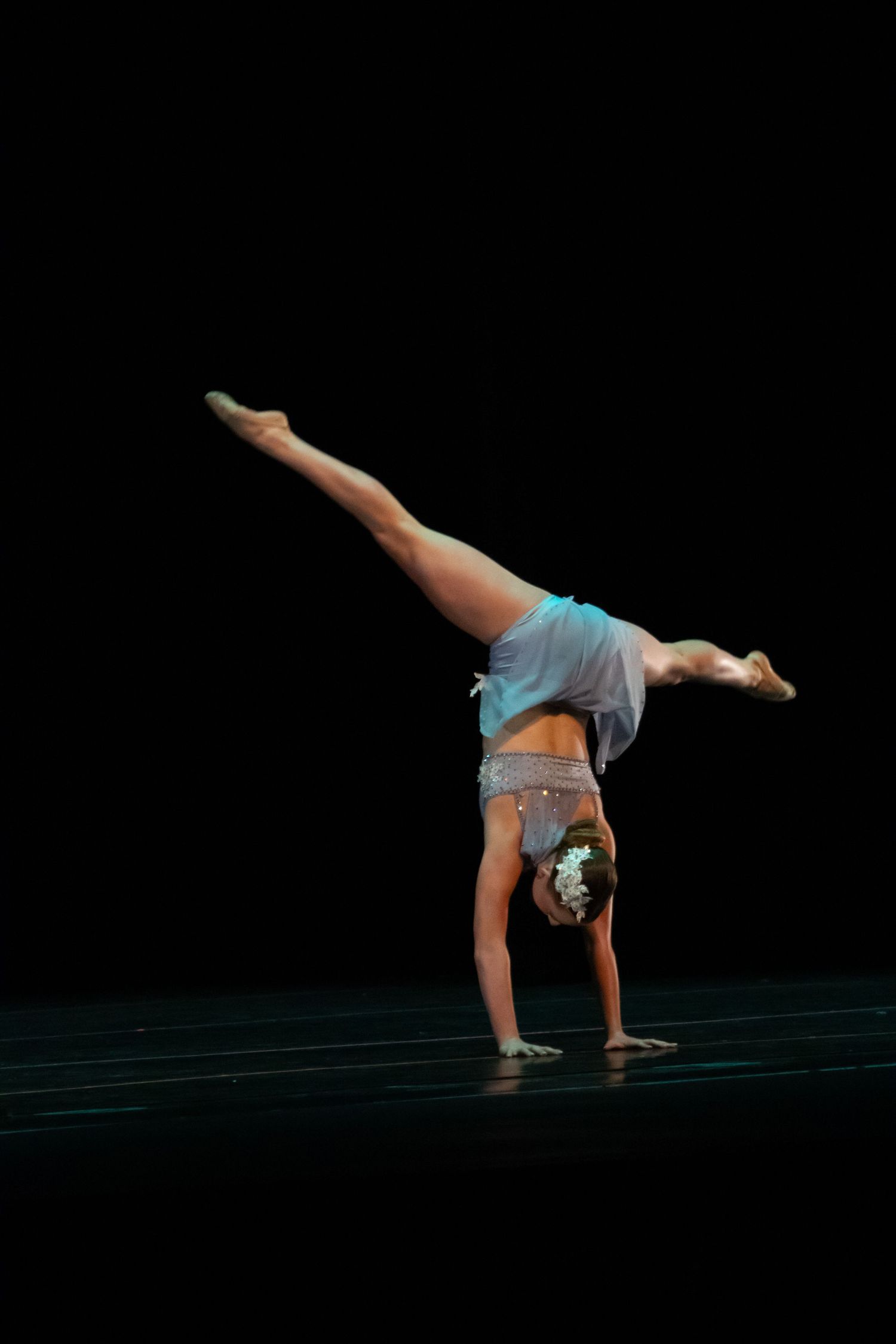 A gymnastic dancer performs a perfect handstand split in a blue costume on a dark stage.