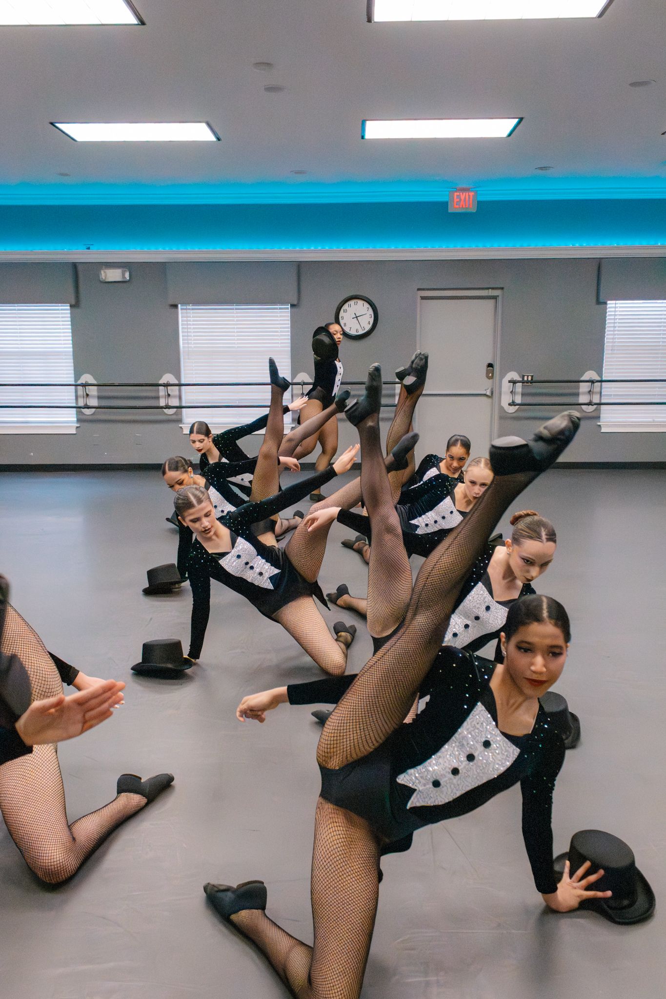 Young dancers in black tuxedo leotards perform a synchronized floor routine with legs raised in a dance studio.