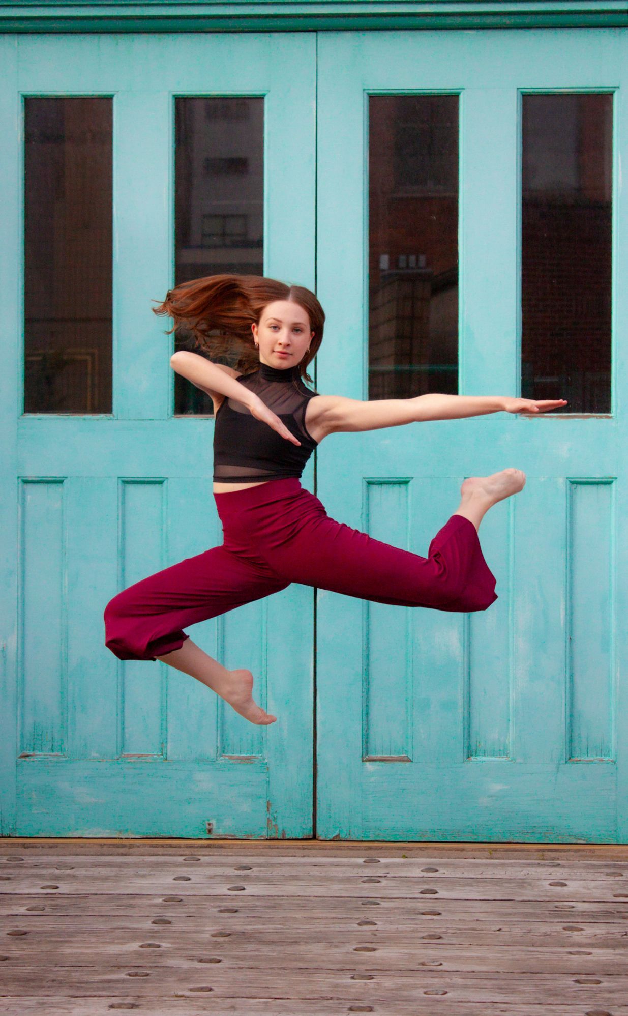 Dynamic female dancer leaping mid-air in red pants before turquoise doors outdoors.