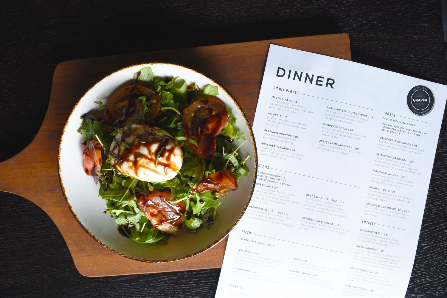 A fresh dinner salad with grilled halloumi, mixed greens, and tomatoes on a wooden board beside a dinner menu.