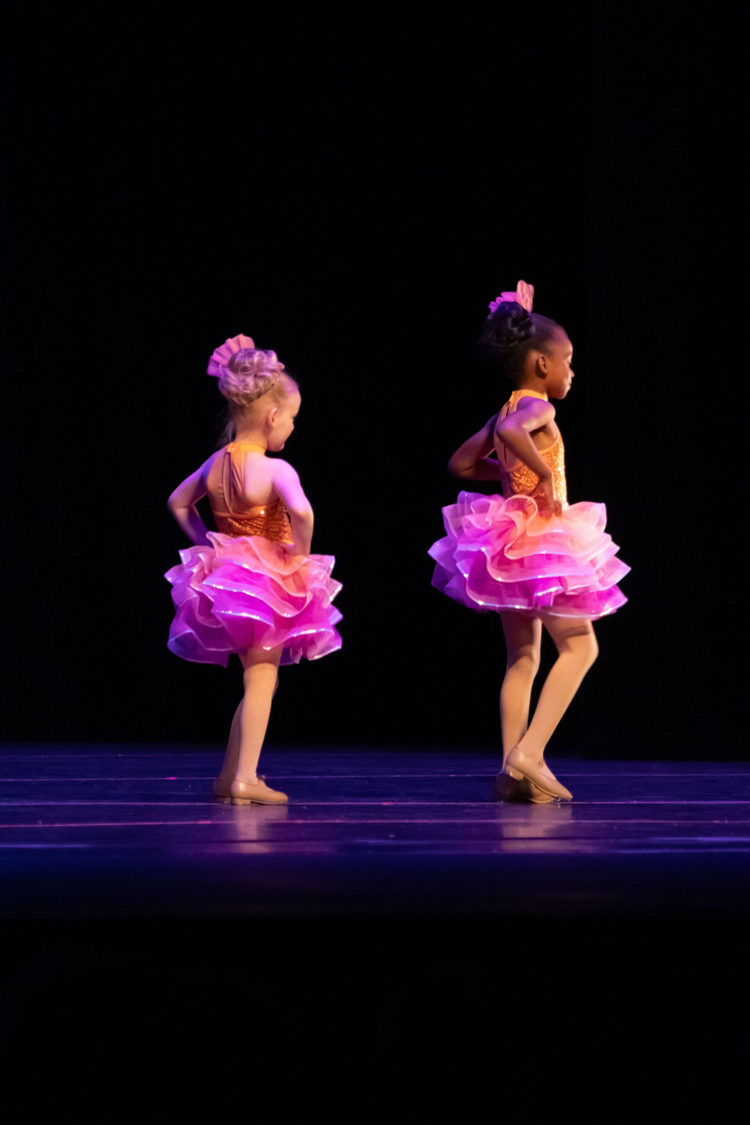 Two young girls in pink ruffled tutus perform ballet on a dark stage during a dance recital.