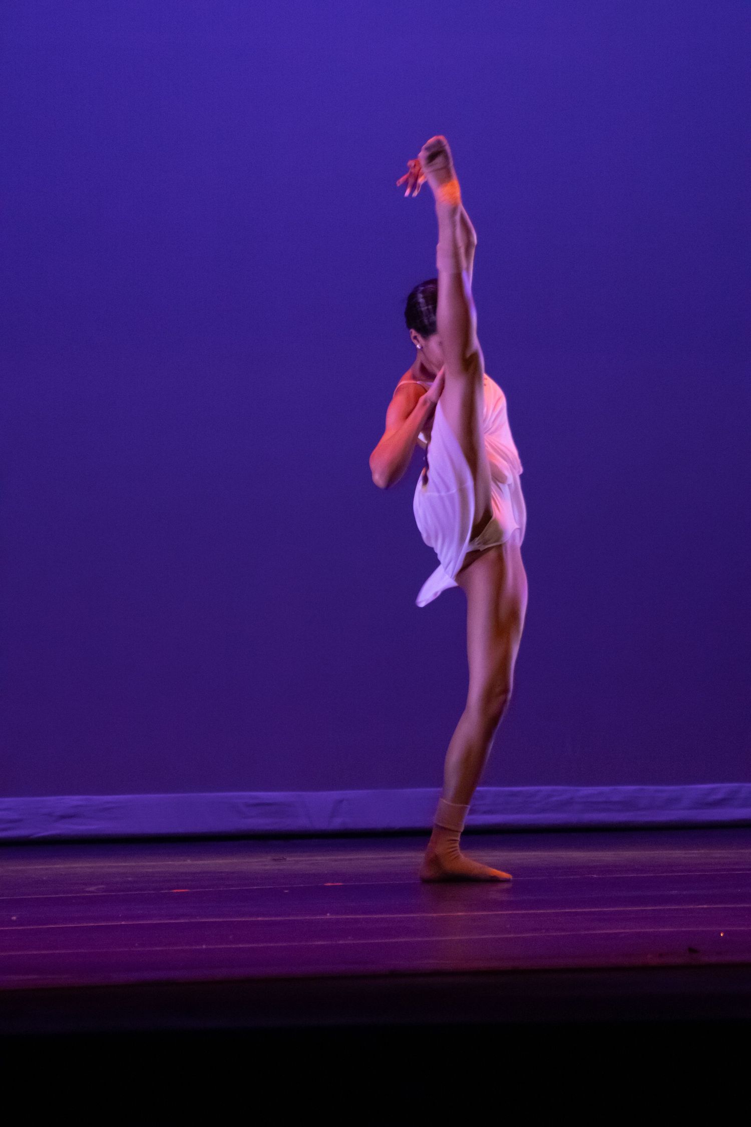 A dancer in a white costume stretches upward in a graceful full-body extension on a purple-lit stage.
