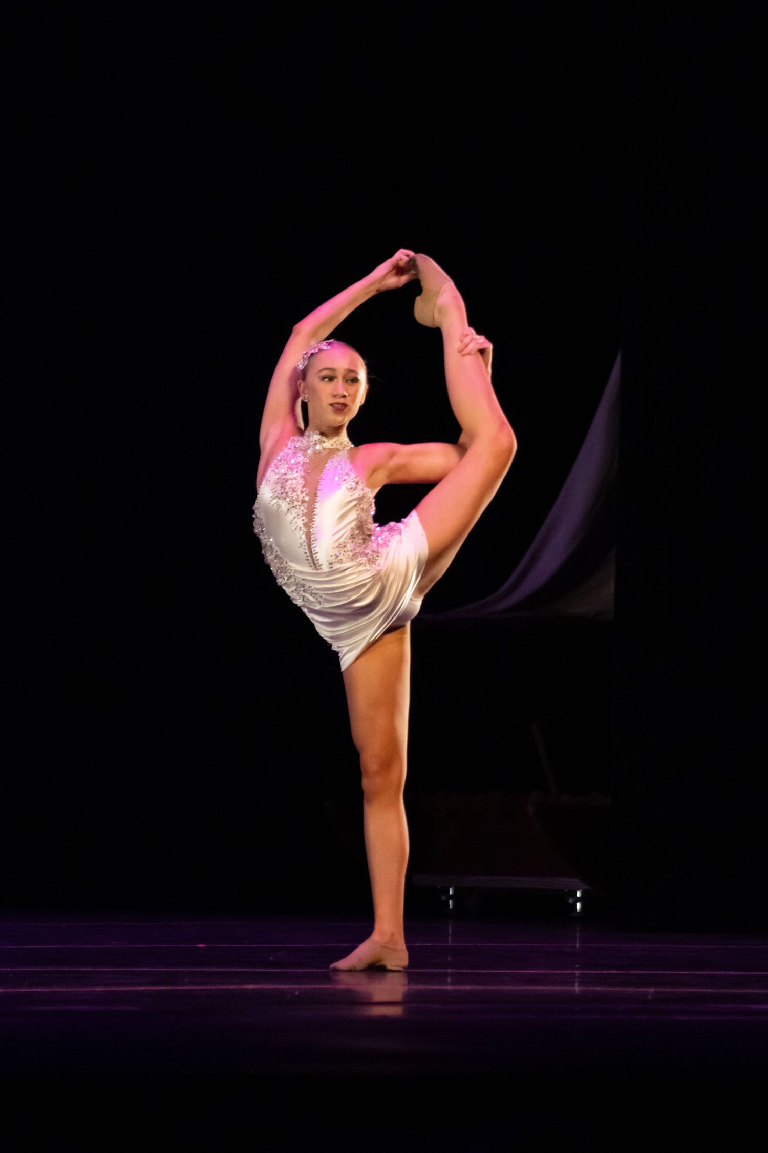 A skilled dancer in a white leotard executes a perfect standing split under pink stage lighting.