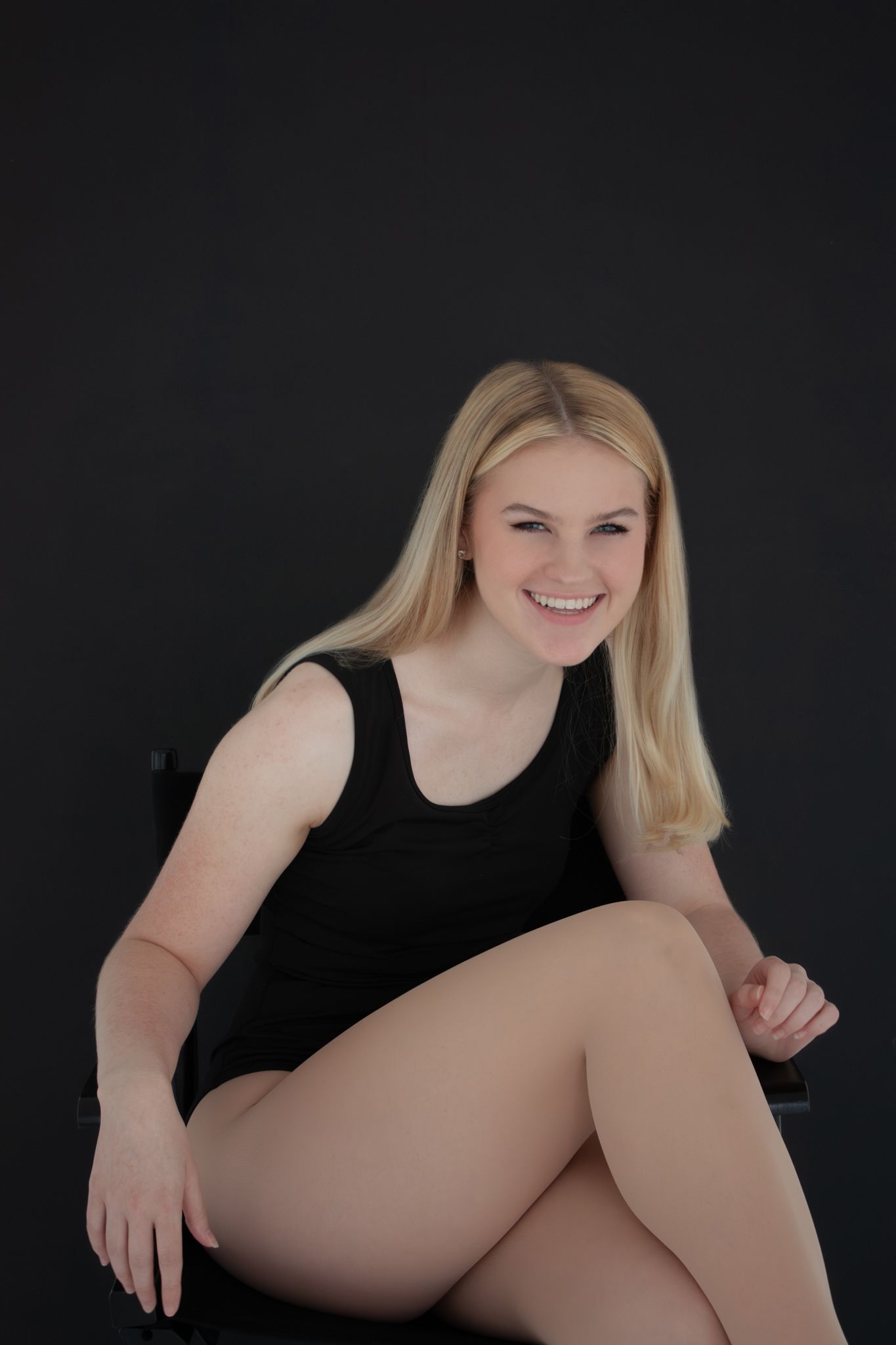 Smiling blonde woman in black tank top sitting cross-legged on chair against dark background.