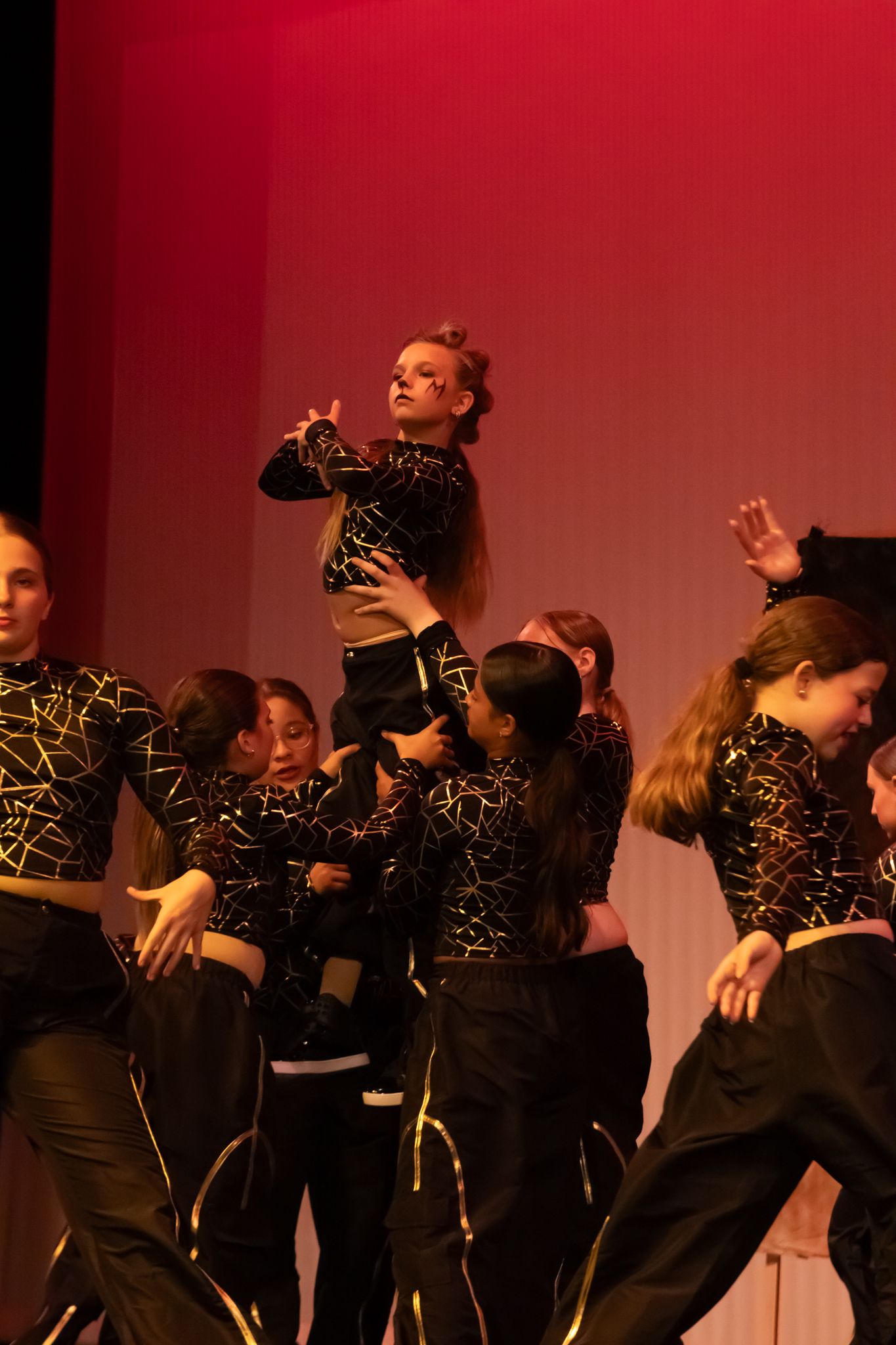 Young dancers in black and gold costumes perform hip-hop routine with one girl lifted on stage.